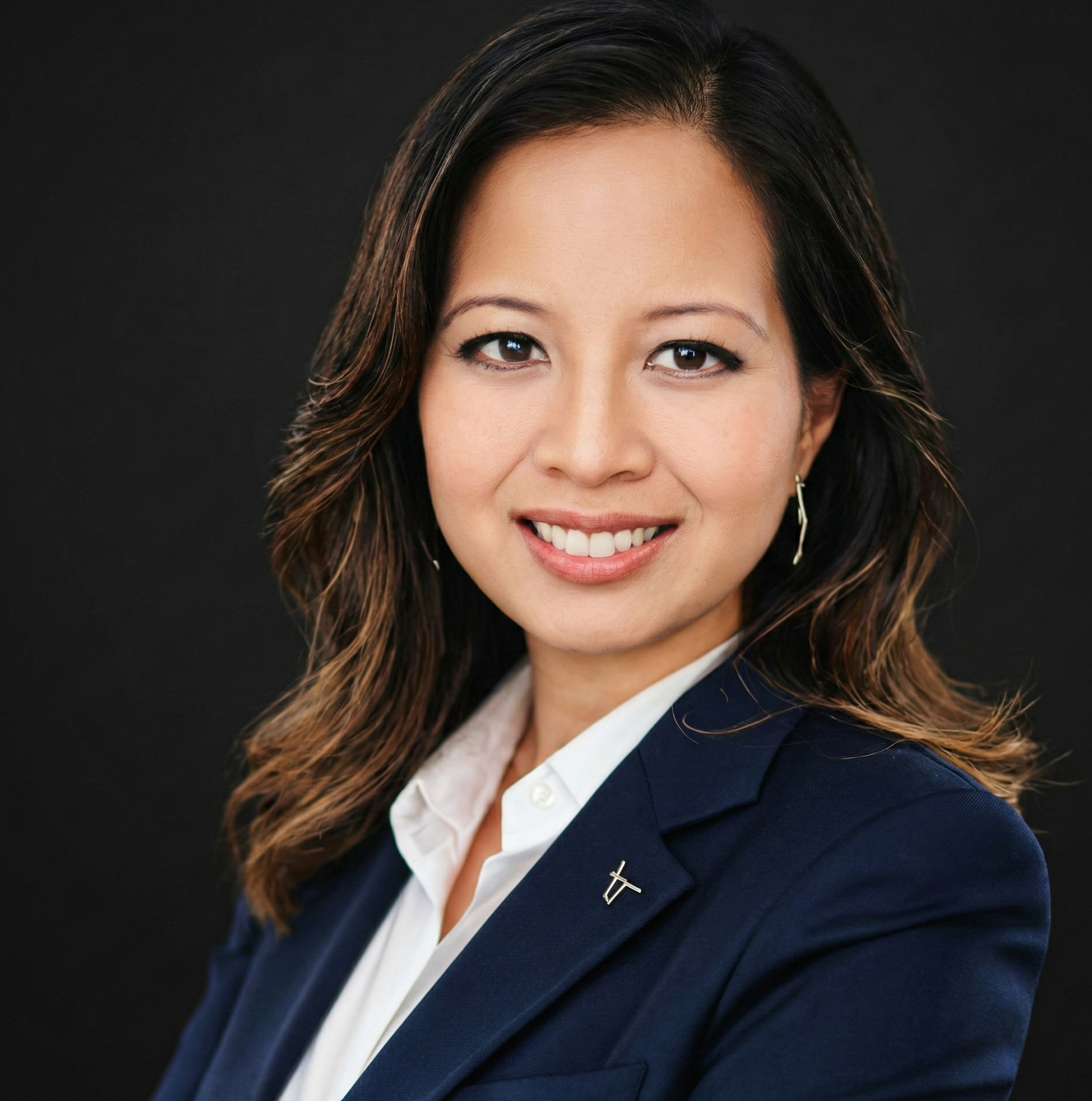A professional headshot of a smiling woman with shoulder-length wavy hair, wearing a dark blazer over a white shirt, against a dark background.