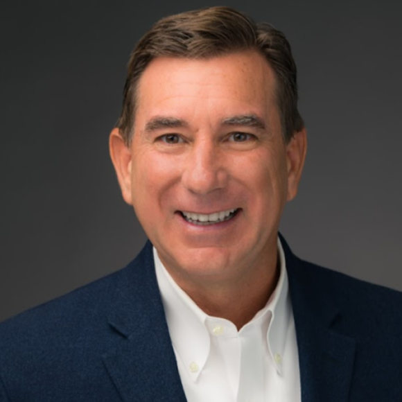 Headshot of a smiling man wearing a dark blazer and white shirt, against a gray background.
