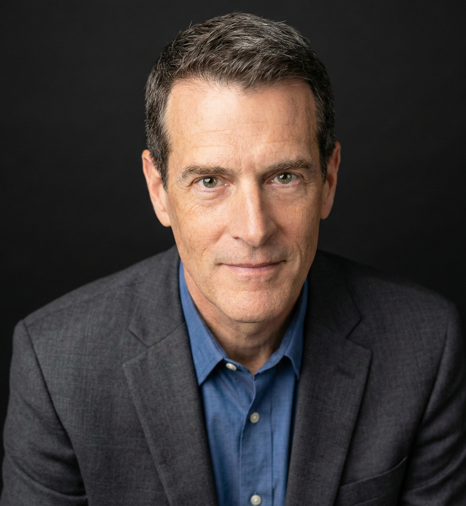 A middle-aged man with short dark hair, green eyes, and light skin, wearing a dark gray suit jacket and a blue dress shirt, smiling subtly against a black background.