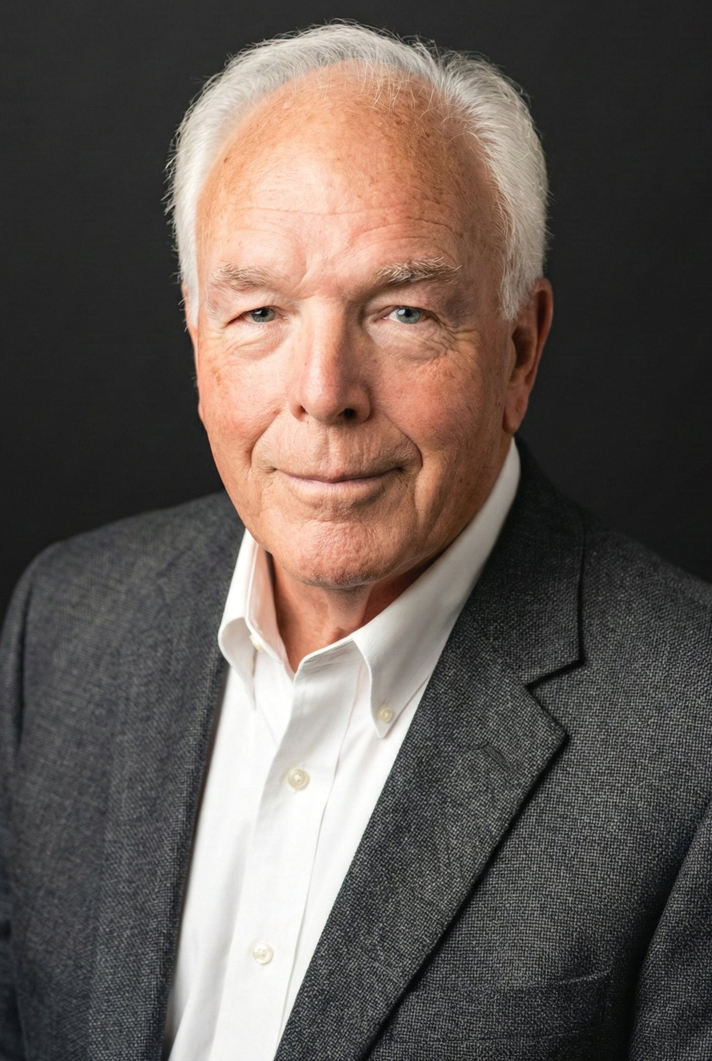 An older man with white hair and light skin, wearing a white shirt and dark gray blazer, posing against a solid dark background.