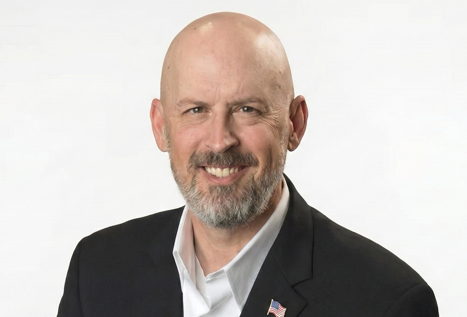 A middle-aged man with a bald head, gray beard, and smiling face, wearing a black suit jacket and white shirt, with a small American flag pin on his lapel, posing against a white background.