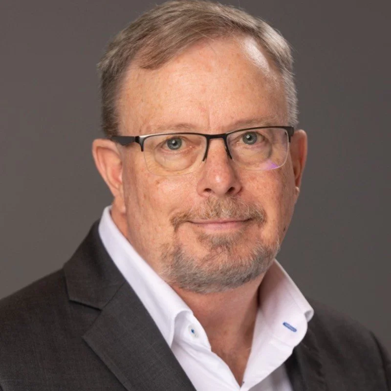 A middle-aged man with short light brown hair, glasses, and a goatee, wearing a dark suit jacket and white dress shirt, posing against a gray background.