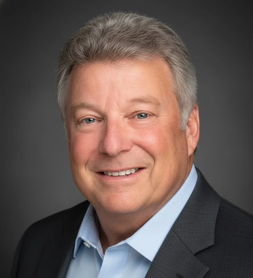 Headshot of a middle-aged man with gray hair, wearing a dark blazer and light blue shirt, smiling against a dark grey background.