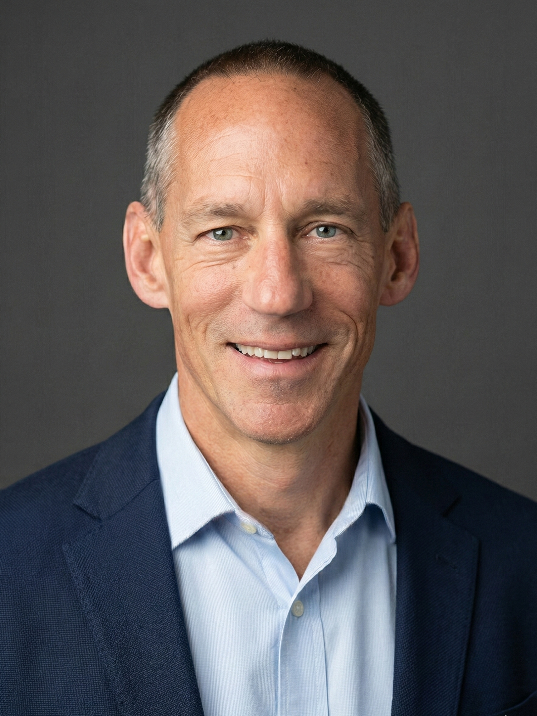 Headshot of a middle-aged man with short brown hair and blue eyes, smiling, wearing a dark blazer and a light blue collared shirt, against a dark gray background.