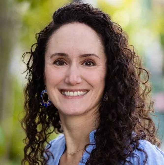 Portrait of a woman with curly brown hair smiling outdoors, wearing blue earrings and a blue top.