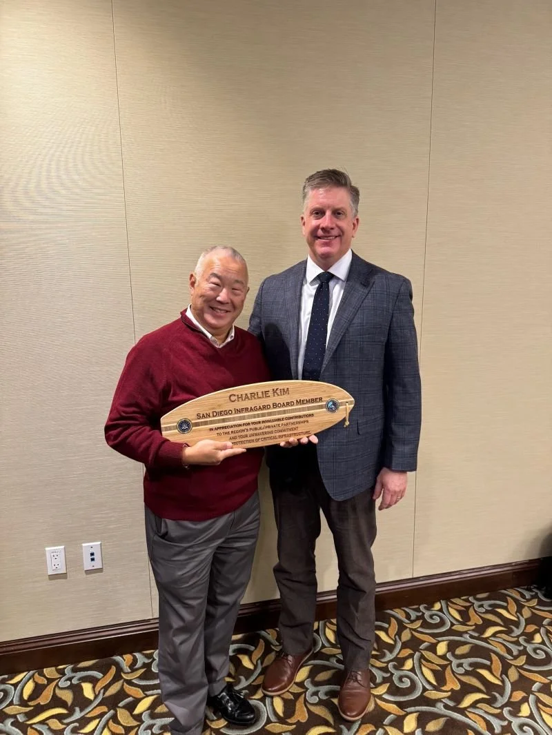 Two men standing indoors, one holding a wooden plaque. The man on the left is wearing a red sweater, gray pants, and black shoes. The man on the right is wearing a dark checkered blazer, white shirt, dark pants, and brown shoes. The plaque has text that reads 'Charlie Kim, San Diego Infragard Board Member.'