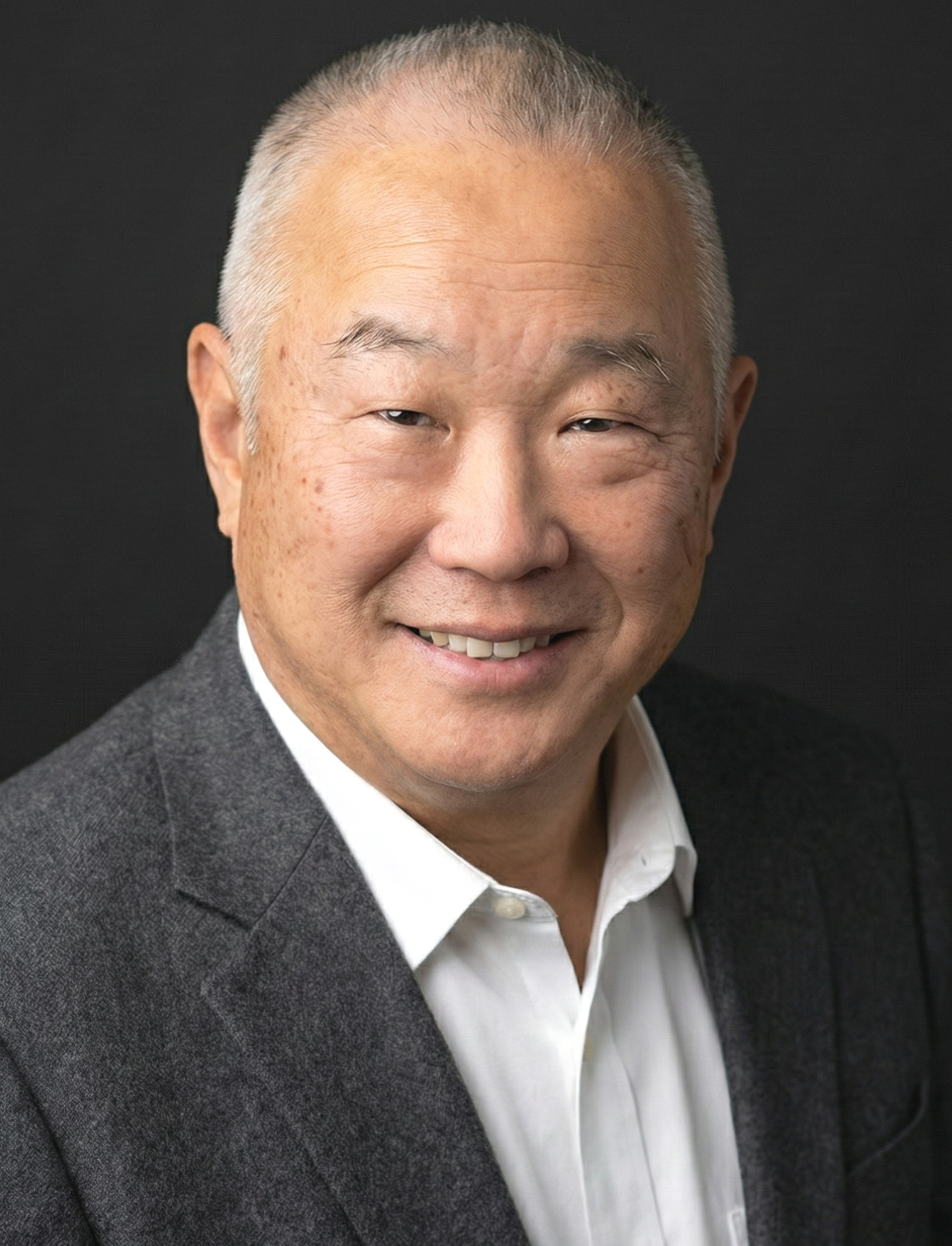 A middle-aged Asian man with short gray hair, smiling, wearing a dark gray blazer and white shirt, against a black background.
