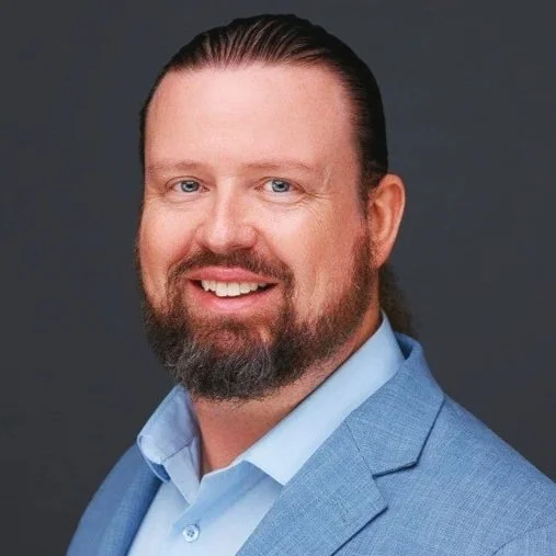Headshot of a smiling man with long hair slicked back, beard, and blue suit jacket, light blue shirt, dark gray background.