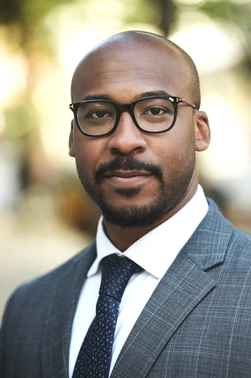 A man wearing glasses, a gray checkered suit, white shirt, and a dark tie, standing outdoors with a blurred background of trees and sunlight.