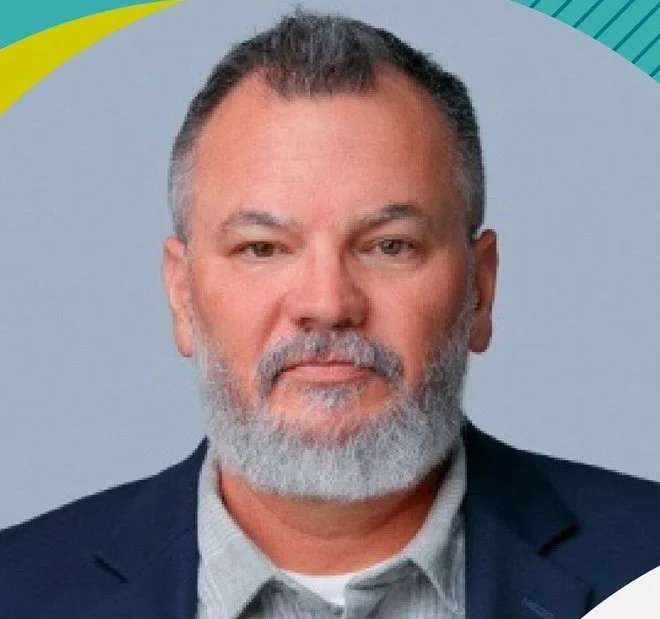 A middle-aged man with a beard and short hair, wearing a dark blazer and a light shirt, against a colorful abstract background.