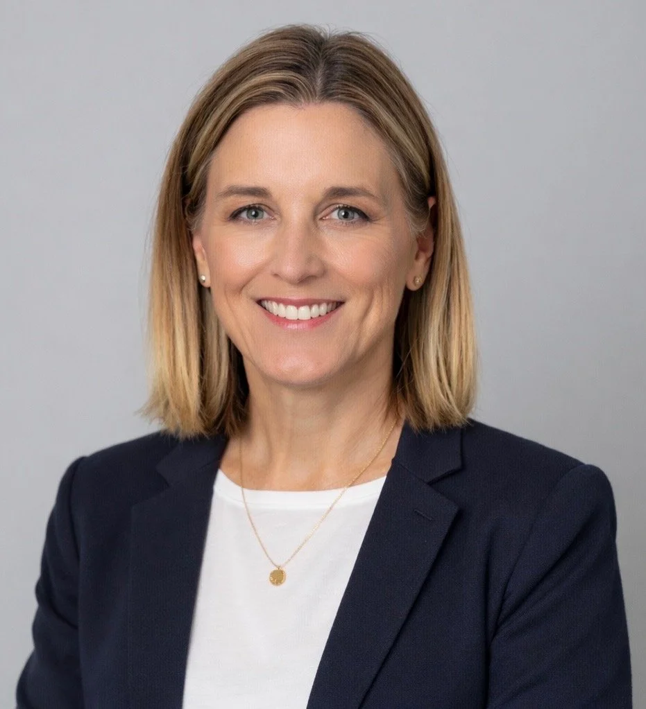 A woman with shoulder-length blonde hair, wearing a dark blazer, white shirt, and a gold necklace, smiling against a plain gray background.