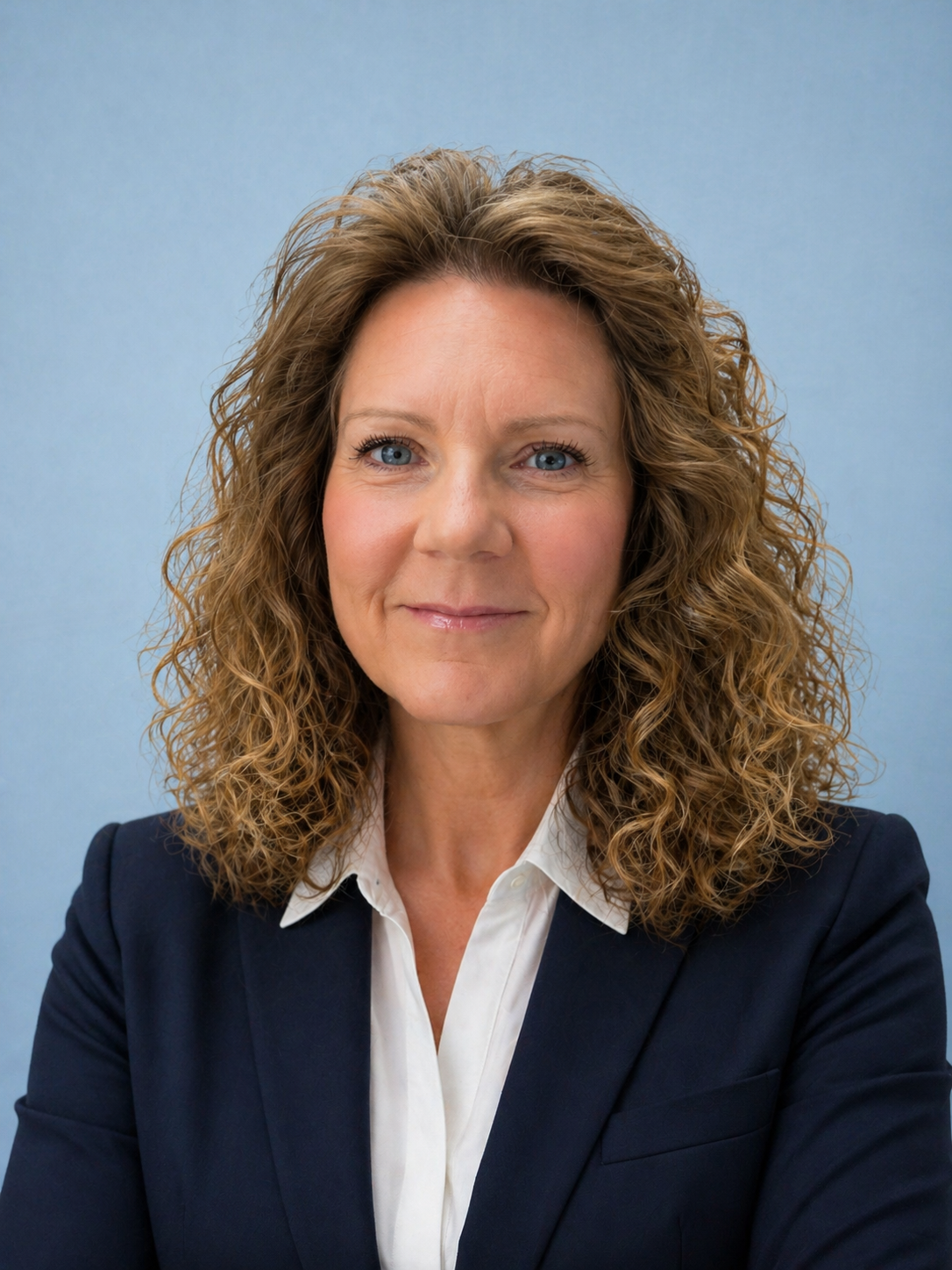 A woman with wavy, shoulder-length light brown hair and blue eyes, wearing a dark blazer over a white shirt, smiling slightly against a light blue background.