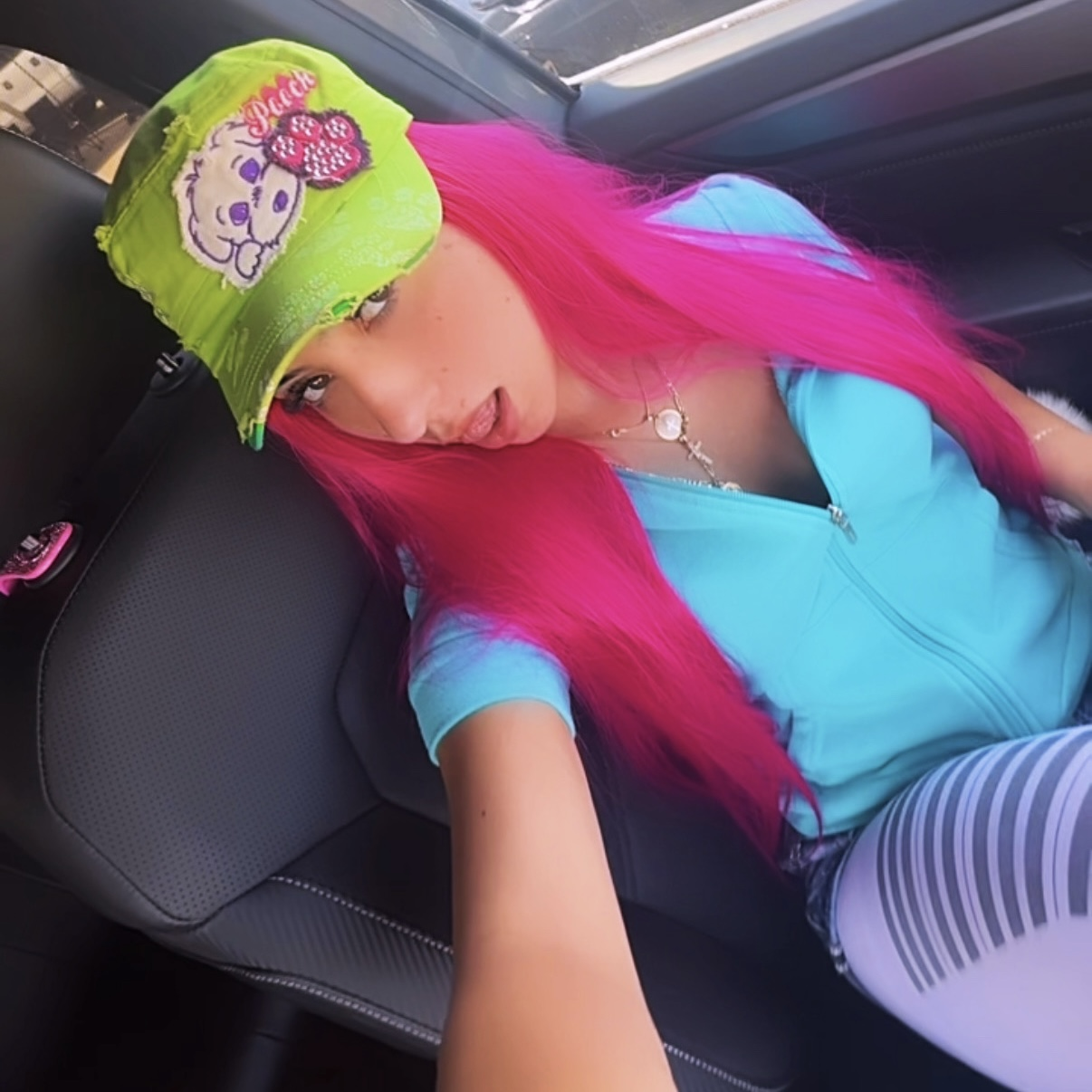 Artist with long pink hair, a lime green cap, teal zip up vest, denim shorts with piano tights posing in a car.