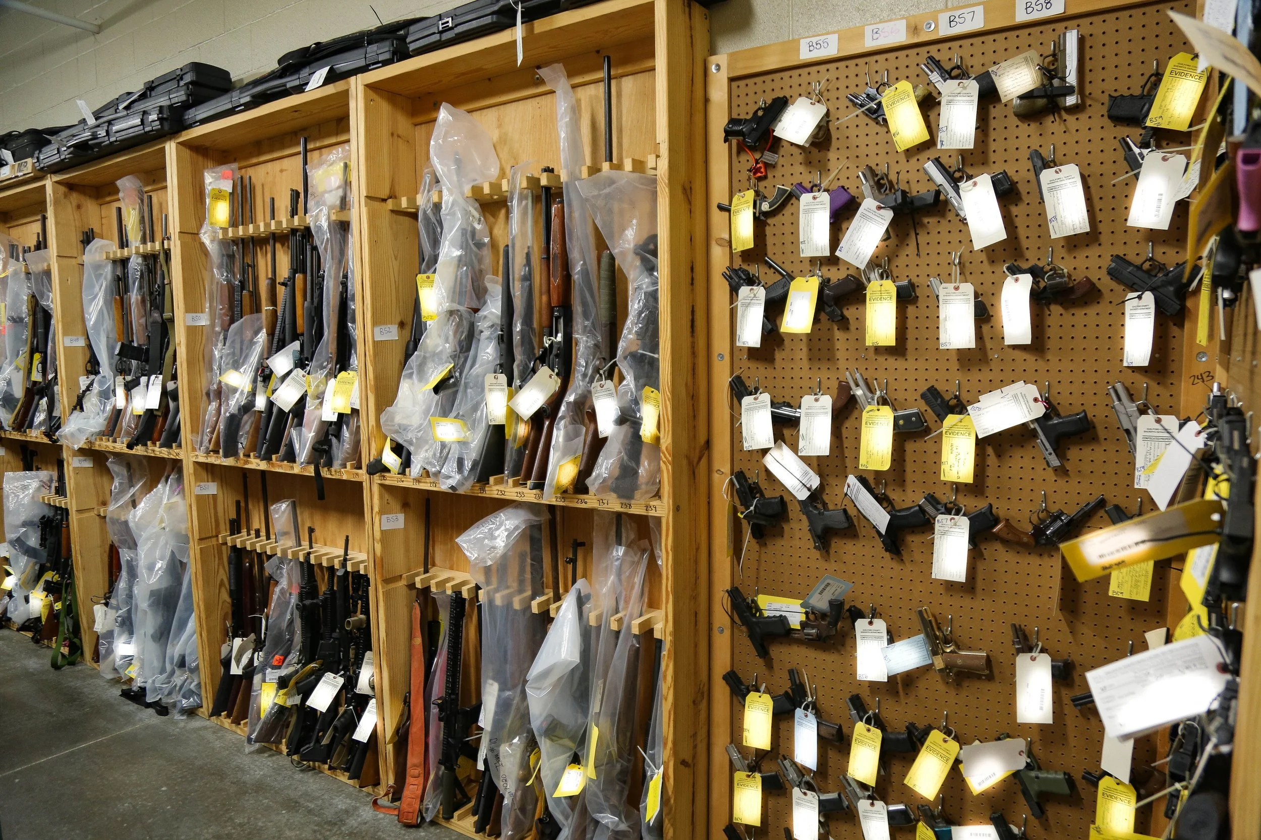 Shelves and a pegboard filled with firearms in a gun store, with some guns in plastic bags on the left and others hanging on the right, each with a yellow watch tag.