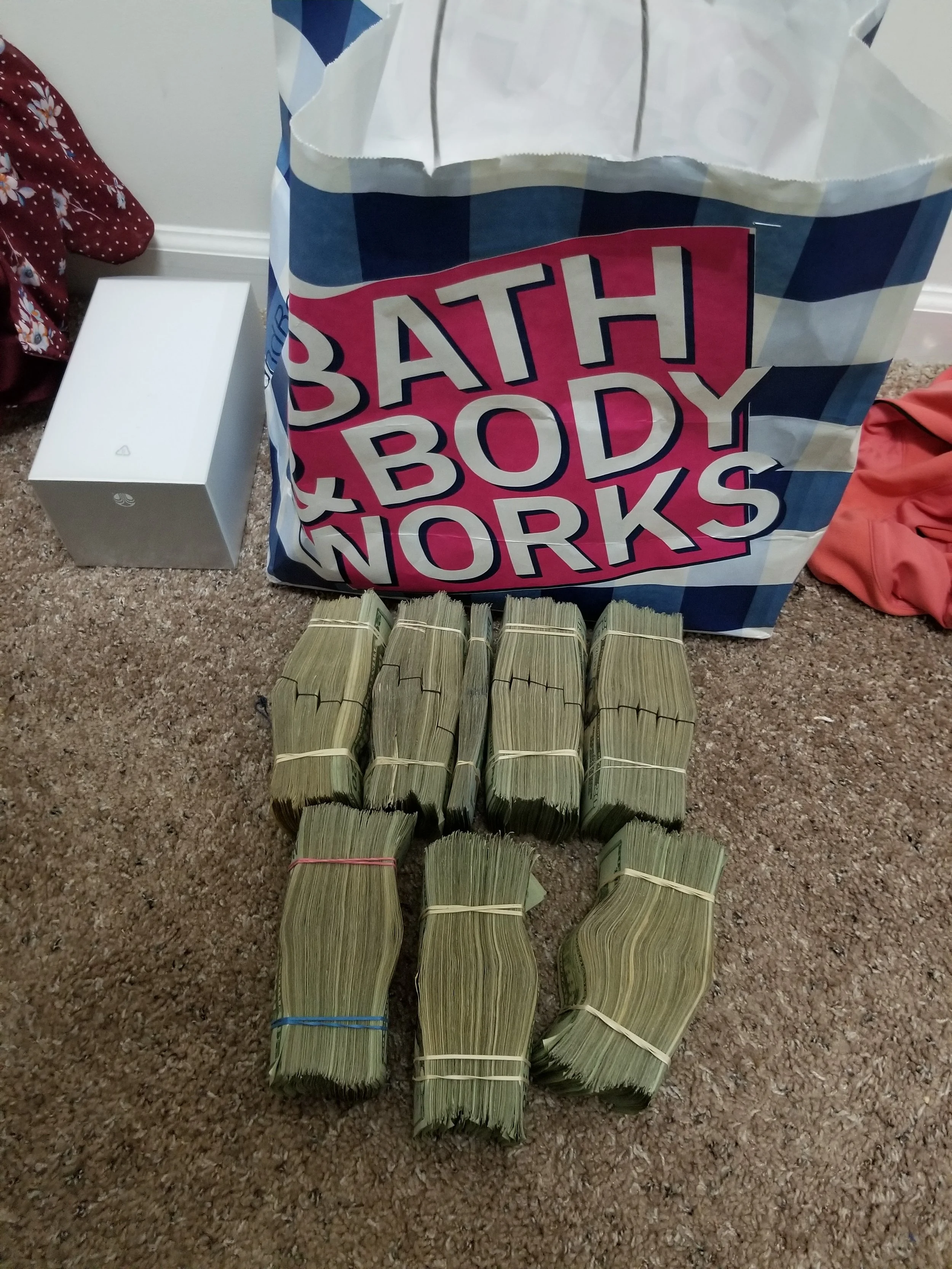Multiple stacks of cash arranged on a beige carpet in front of a Bath & Body Works shopping bag. To the left, there is a white box and some clothing items around.