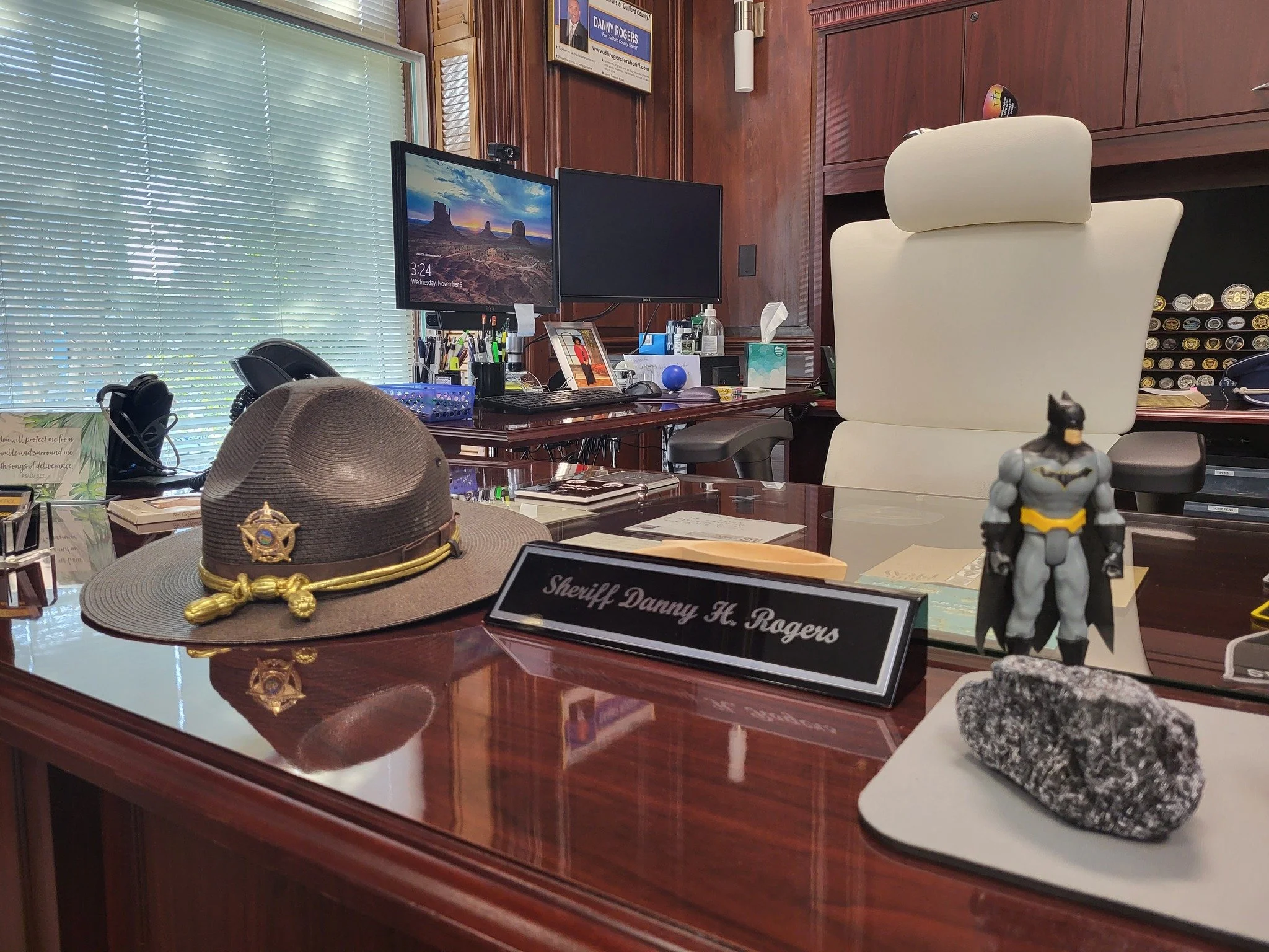 An office desk with a sheriff's hat, a Batman figurine, a nameplate reading 'Sheriff Danny H. Rogers,' and various office supplies. There are two computer monitors and a white office chair behind the desk. The background shows wood-paneled walls, a window with blinds, and framed pictures on the wall.