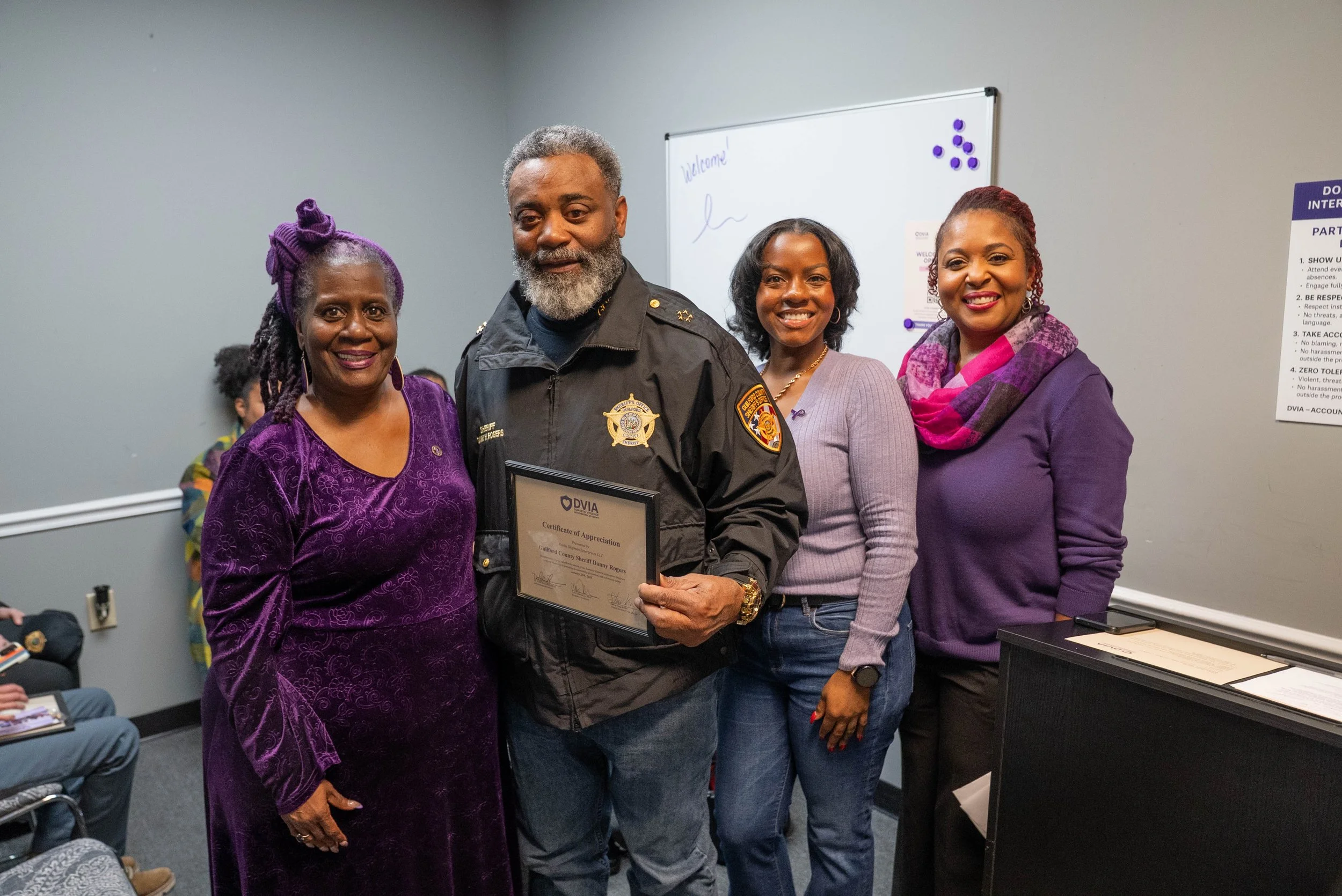 A group of four people, including a police officer holding a framed certificate, standing together in a room, smiling at the camera.
