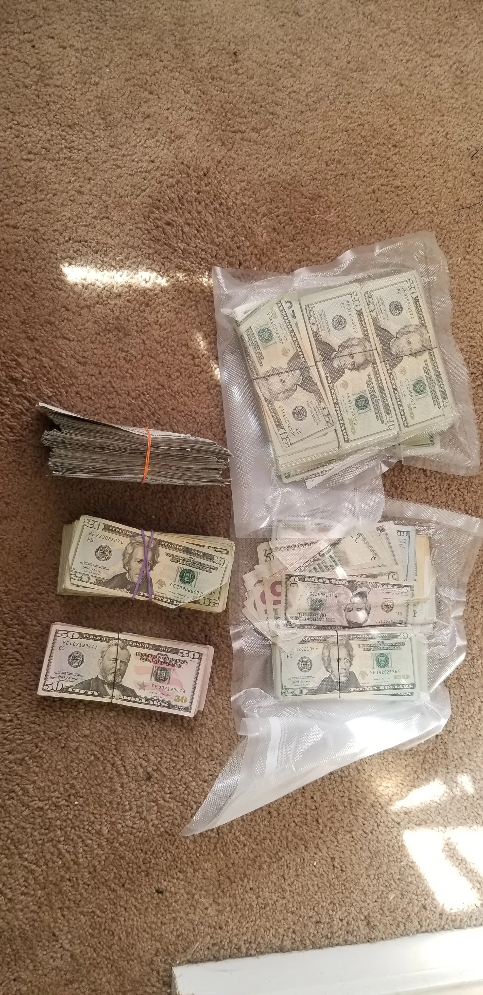Various stacks of U.S. dollar bills, including $20, $50, and $100 denominations, some bundled with rubber bands or in plastic bags, placed on a beige carpet.