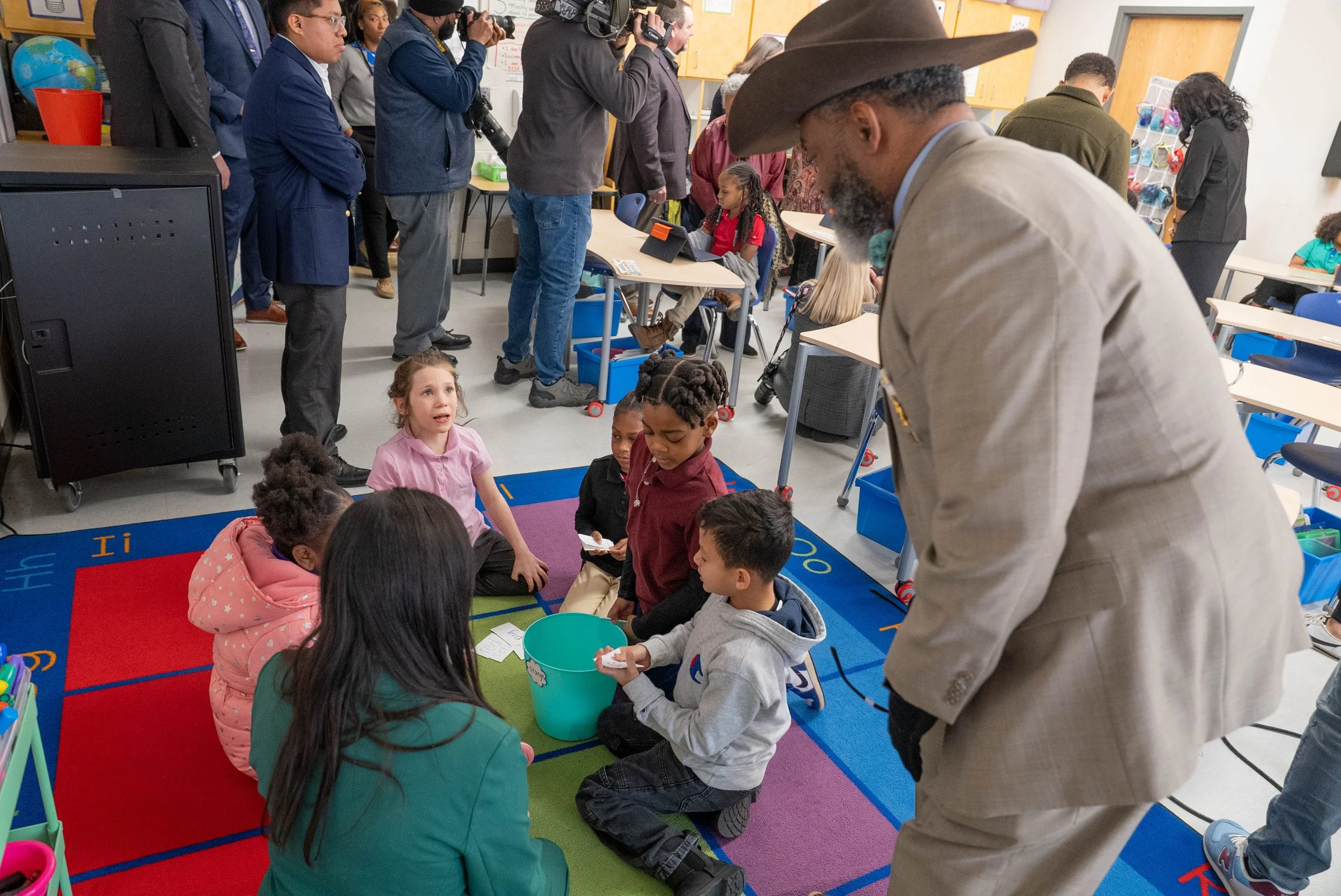 Children sitting on a colorful rug in a classroom, engaging with two adults, one in a beige suit and one in a green blazer. A group of people, including photographers and adults, are observing from behind, with desks and classroom supplies visible in