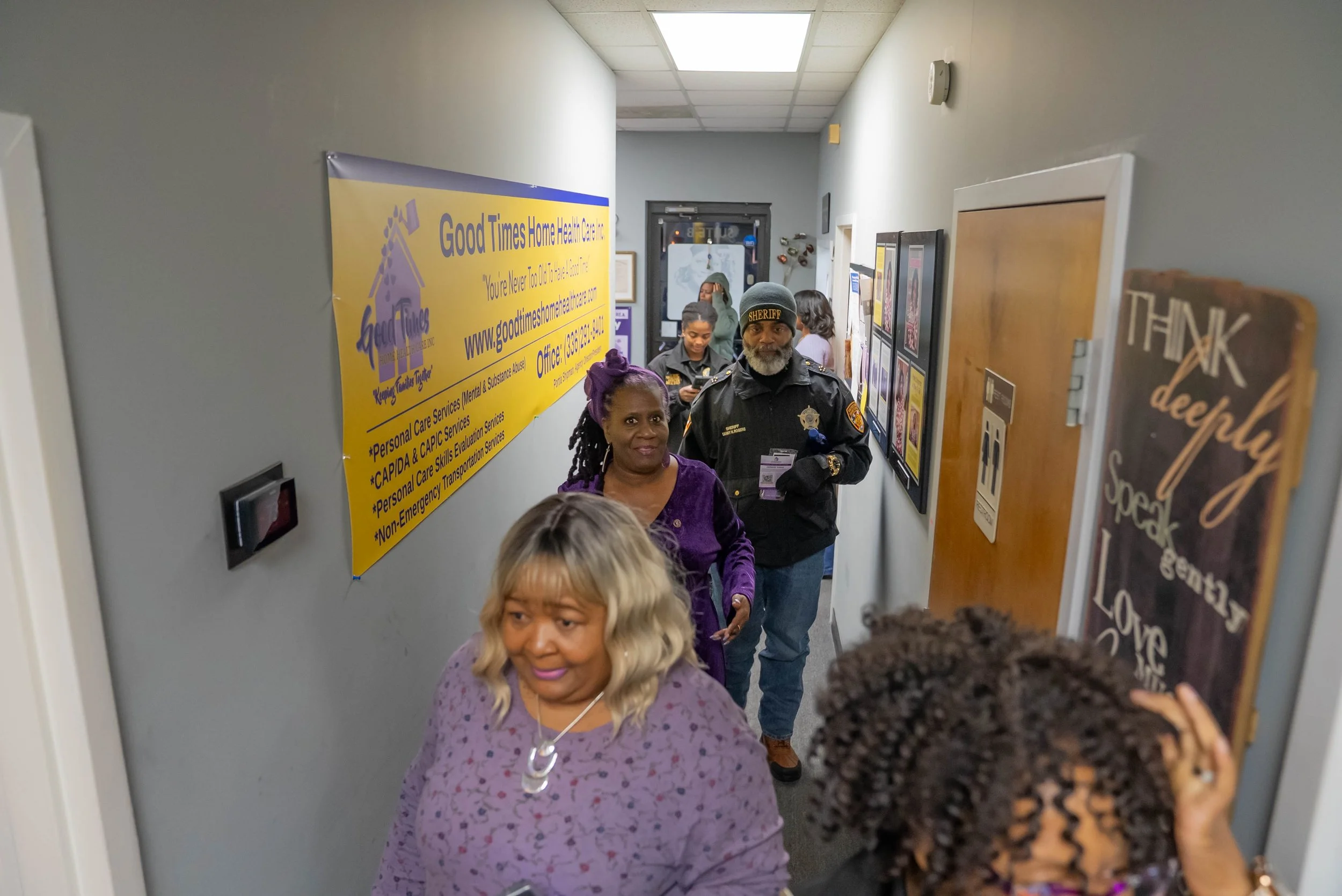 People walking through a hallway at Good Times Home Health Care office, which has a yellow sign on the wall, bulletin boards, and a decorative chalkboard sign.