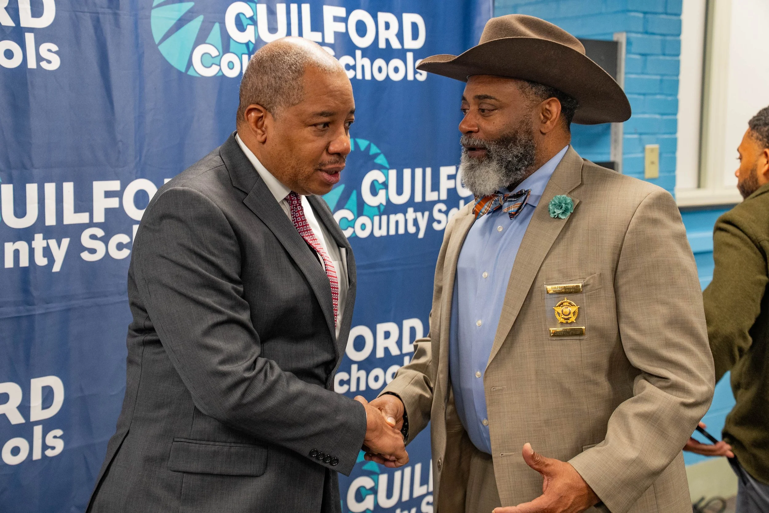 Two men in suits shaking hands at an event with a blue backdrop that reads 'Guilford County Schools'.