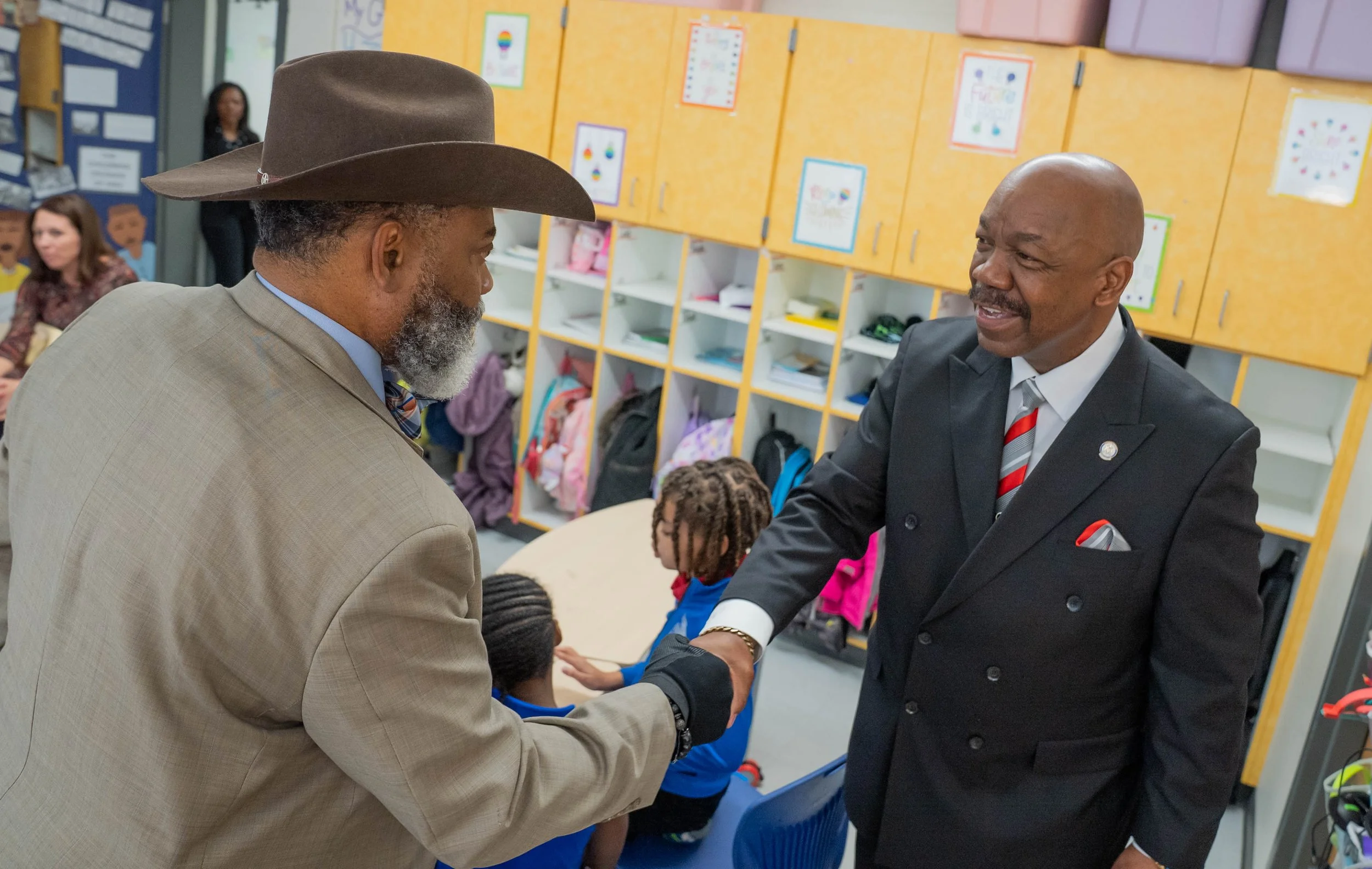 Two men shaking hands in a classroom, one wearing a suit and hat, the other in a suit and tie, with children and lockers in the background.