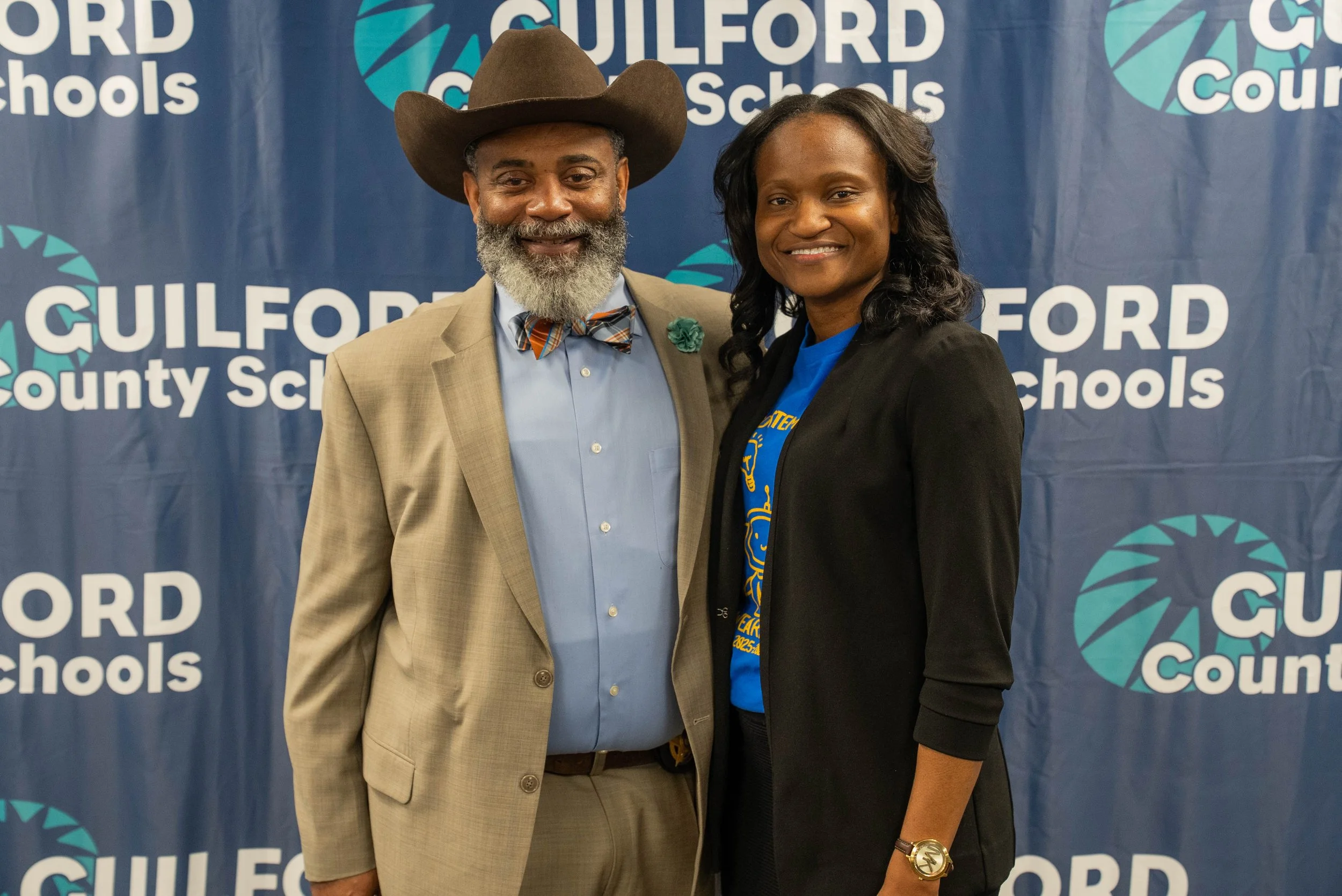 A man and woman standing side by side facing the camera, smiling, with a blue background featuring 'Guilford County Schools' logo and text.
