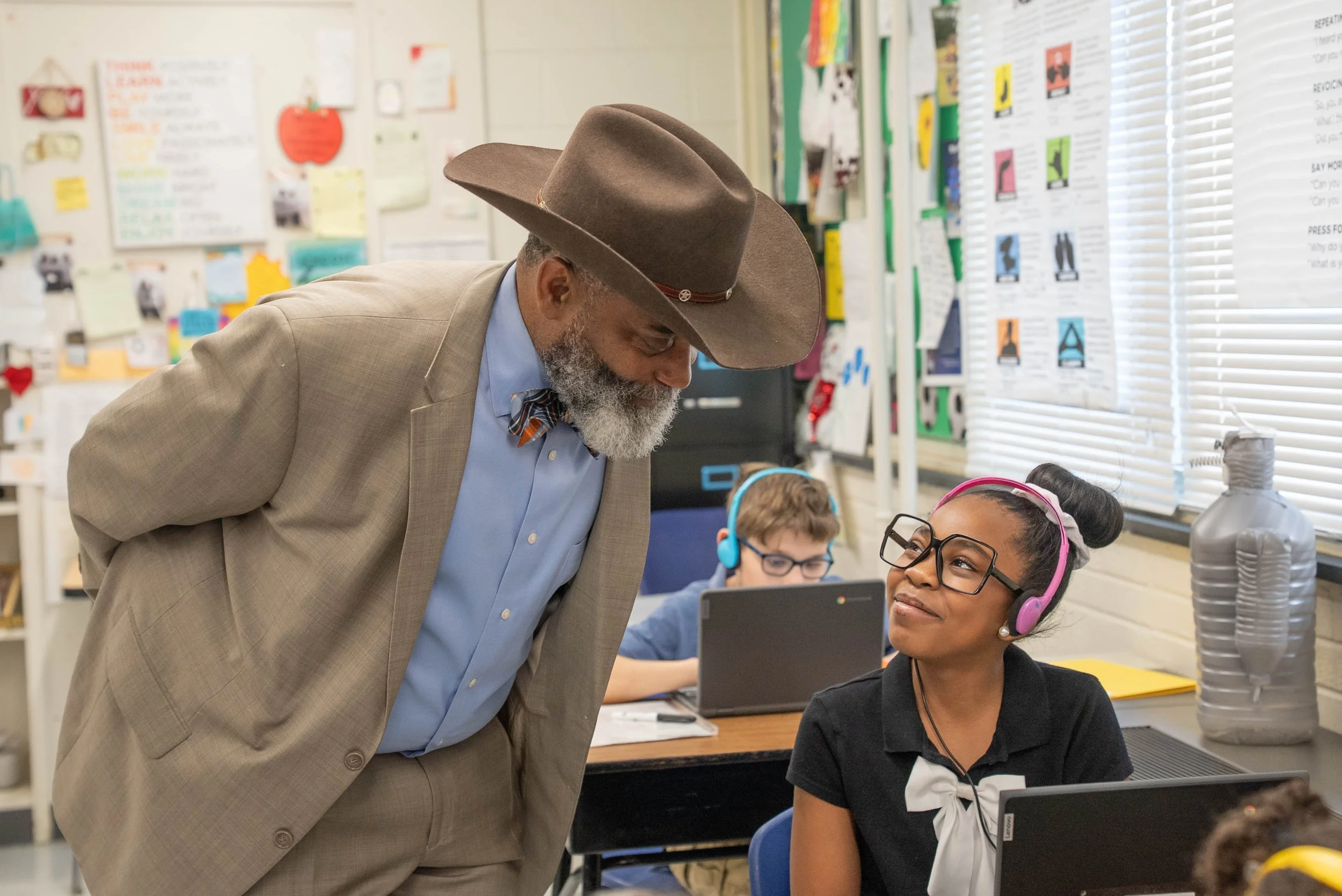 A classroom with students working at computers. An adult man in a brown suit and cowboy hat is leaning over to speak with an African American girl with glasses and pink headphones, who is looking up at him. A boy with glasses and blue headphones is working on a laptop in the background.