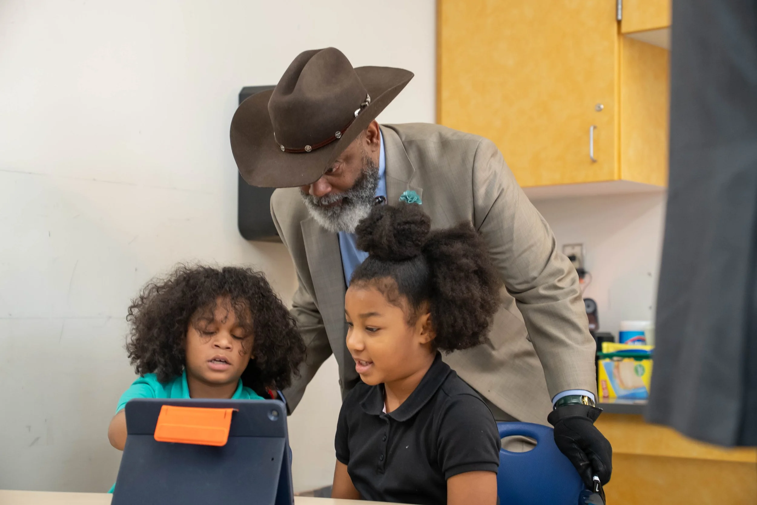 An older man with a gray beard and a cowboy hat looks down and talks to two young girls sitting at a table, one with curly hair and the other with a large bun, while they look at a tablet.