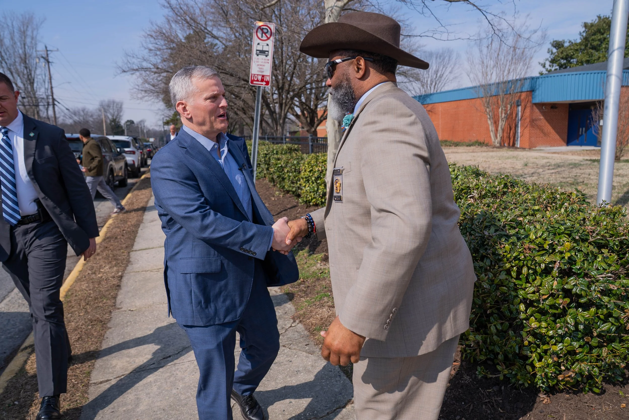 Two men in suits shaking hands outside on a sidewalk near a parking lot and bushes, with other people in the background, and a no parking sign visible.