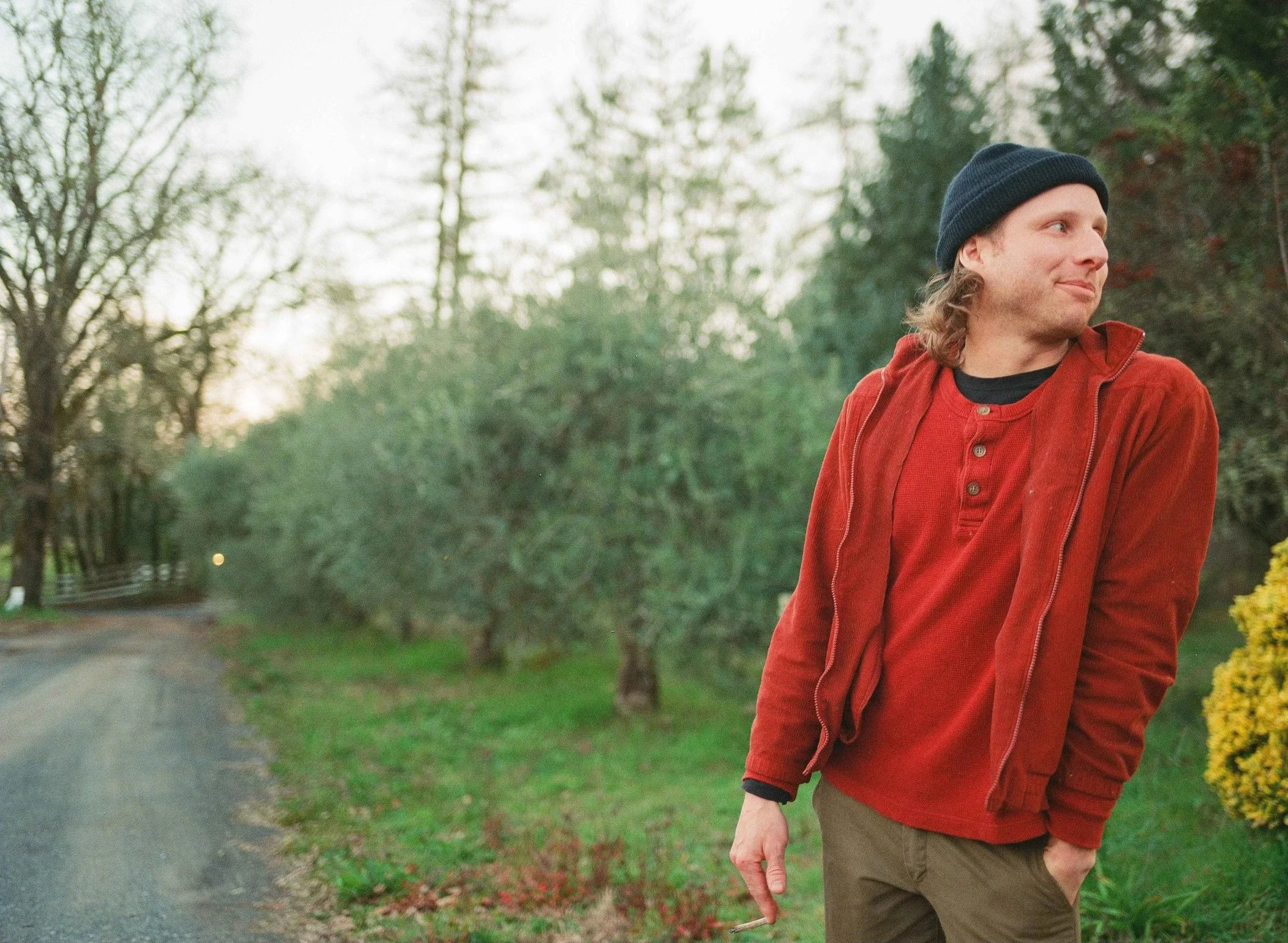 Muunjuun with long hair wearing a dark beanie, red jacket, and khaki pants, standing outdoors on a walk in a park or garden during daytime, with trees and bushes in the background.