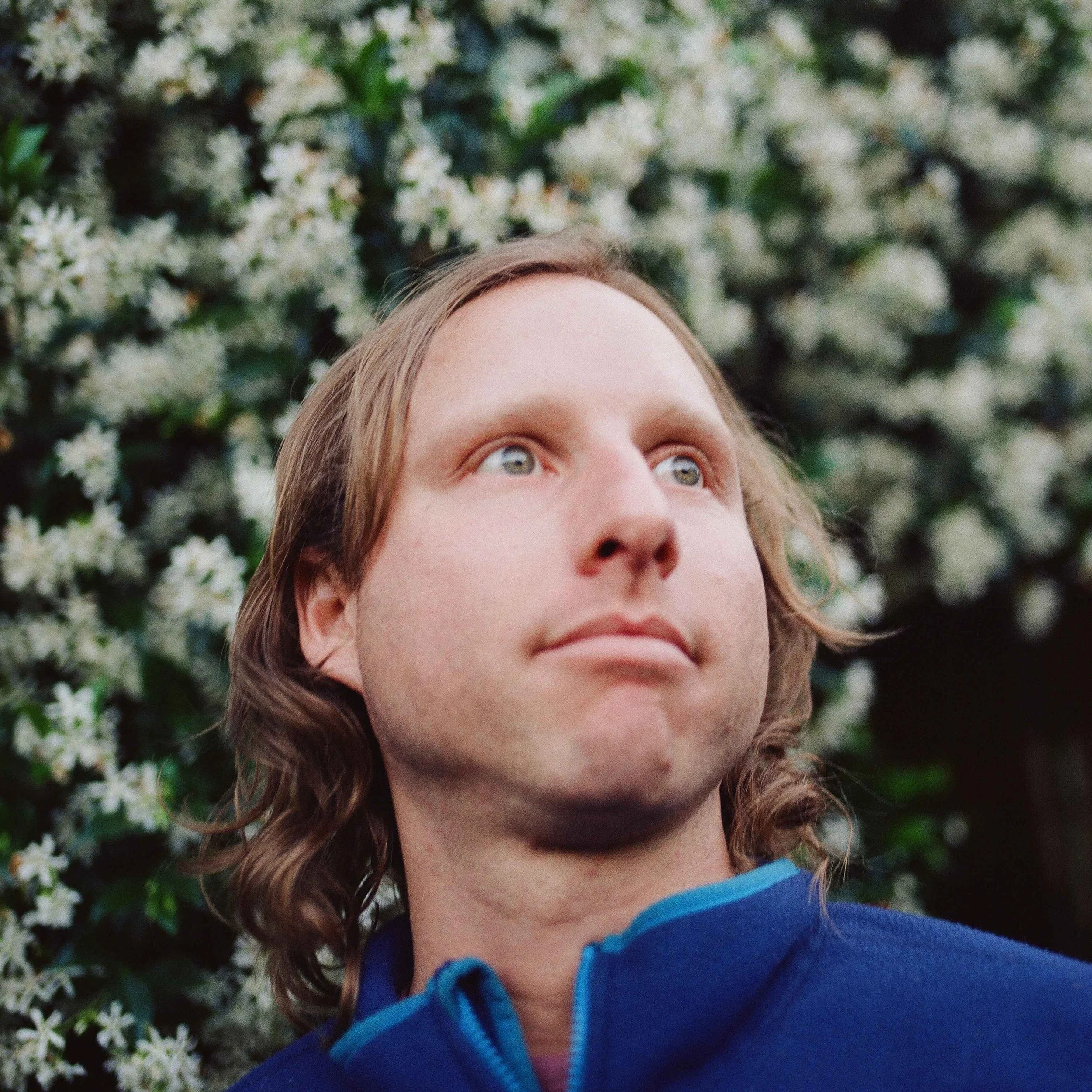 A man with shoulder-length wavy hair looking up and to the right, wearing a blue jacket, with white flowering bushes in the background.