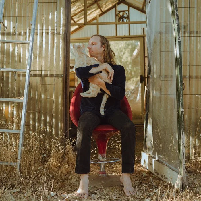 Muunjuun sitting barefoot on a red chair inside a rustic wooden shed, holding and cuddling a big orange tabby cat, with hay and sunlight outside.