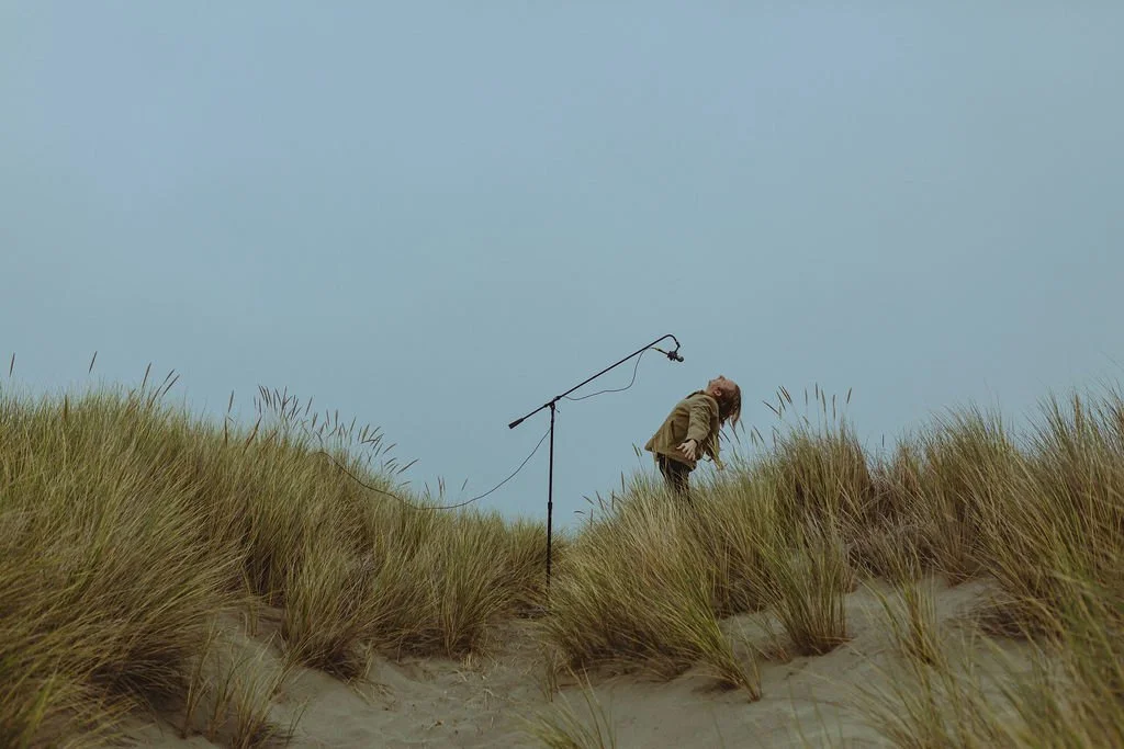 Muunjuun with long hair leaning forward on sandy dune, standing next to a microphone stand with a microphone, under a cloudy sky.