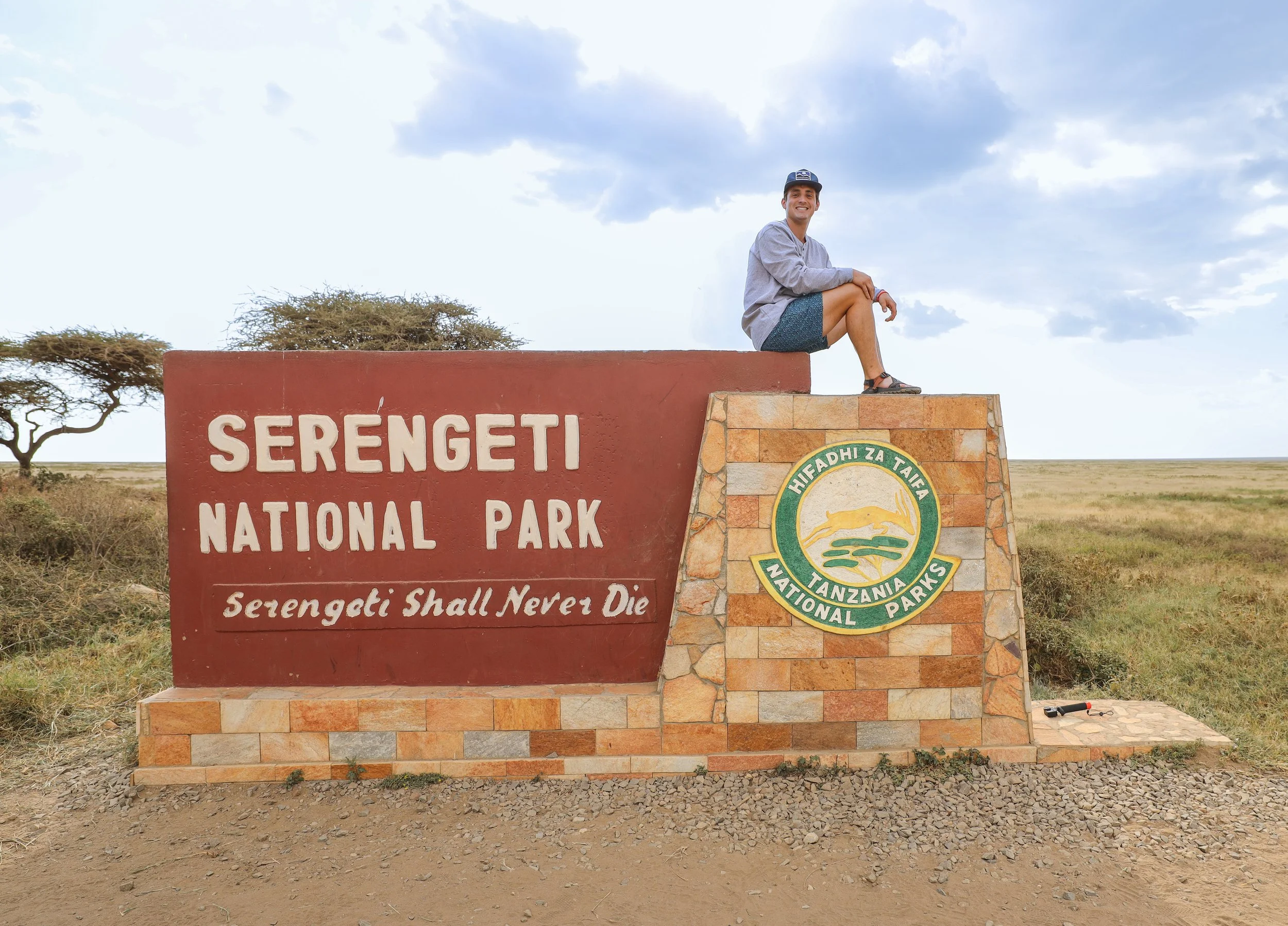 Sebastian sitting on a monument sign at Serengeti National Park in Tanzania, with a kangaroo illustration on a badge, and the phrase 'Serengeti Shall Never Die' on the sign.