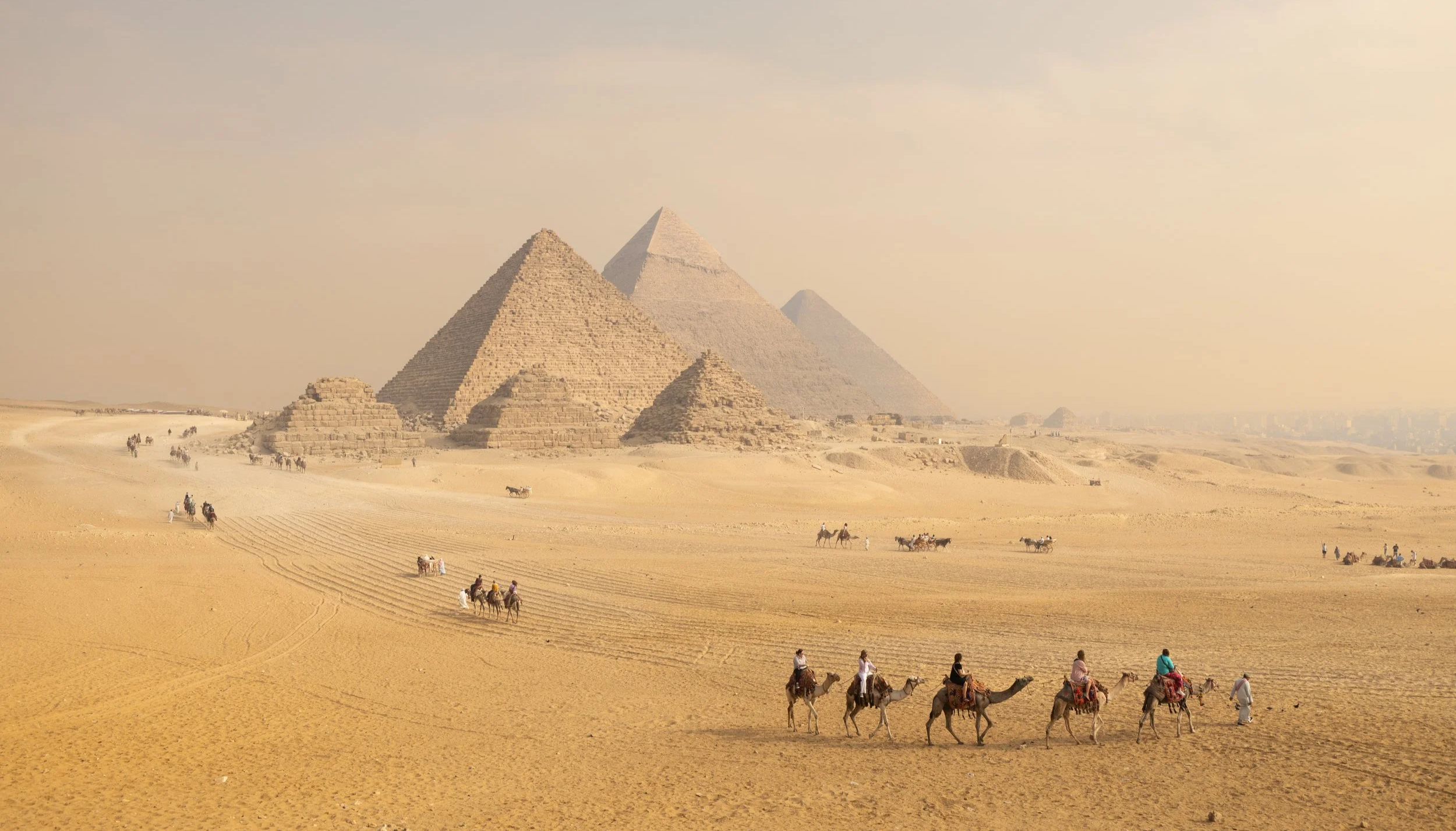 Camels and riders crossing the desert in front of the Pyramids of Giza under a hazy Cairo sky in Egypt.