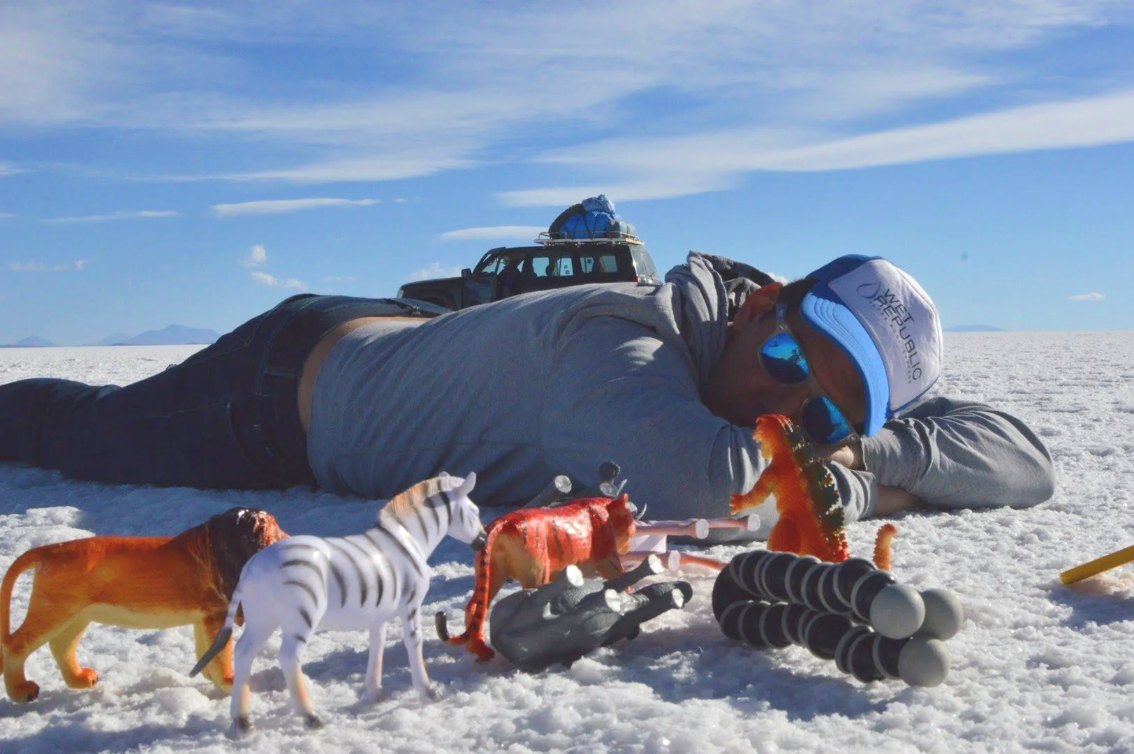 Person lying on salt flats with toy animals and equipment around him, bright sky with some clouds, vehicle in the background.