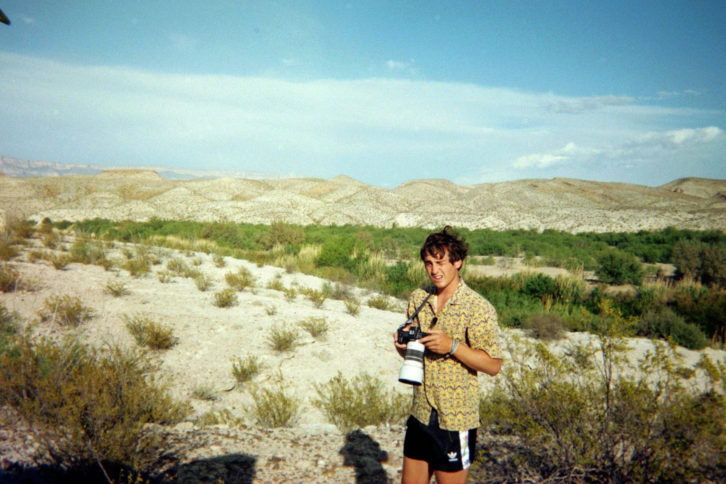 Sebastian in a patterned shirt and black shorts stands in a desert landscape holding a camera with a large telephoto lens, with mountains and sparse vegetation in the background.