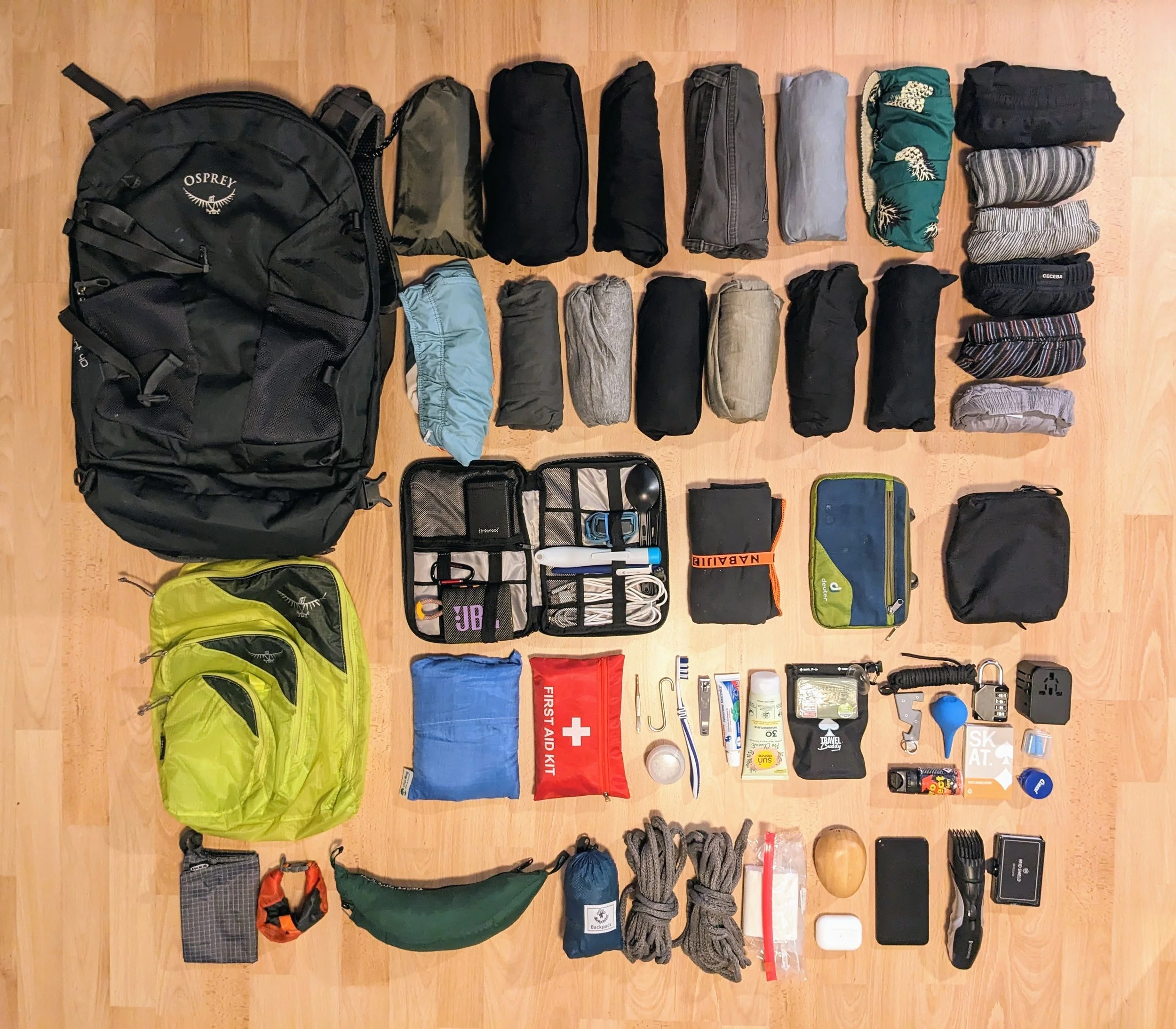 A variety of travel and outdoor gear including backpacks, clothing, toiletries, and accessories organized on a wooden floor. Items include a large black Osprey backpack, smaller bags, clothing items, a first aid kit, toiletries, electronic devices, ropes, and other travel essentials.