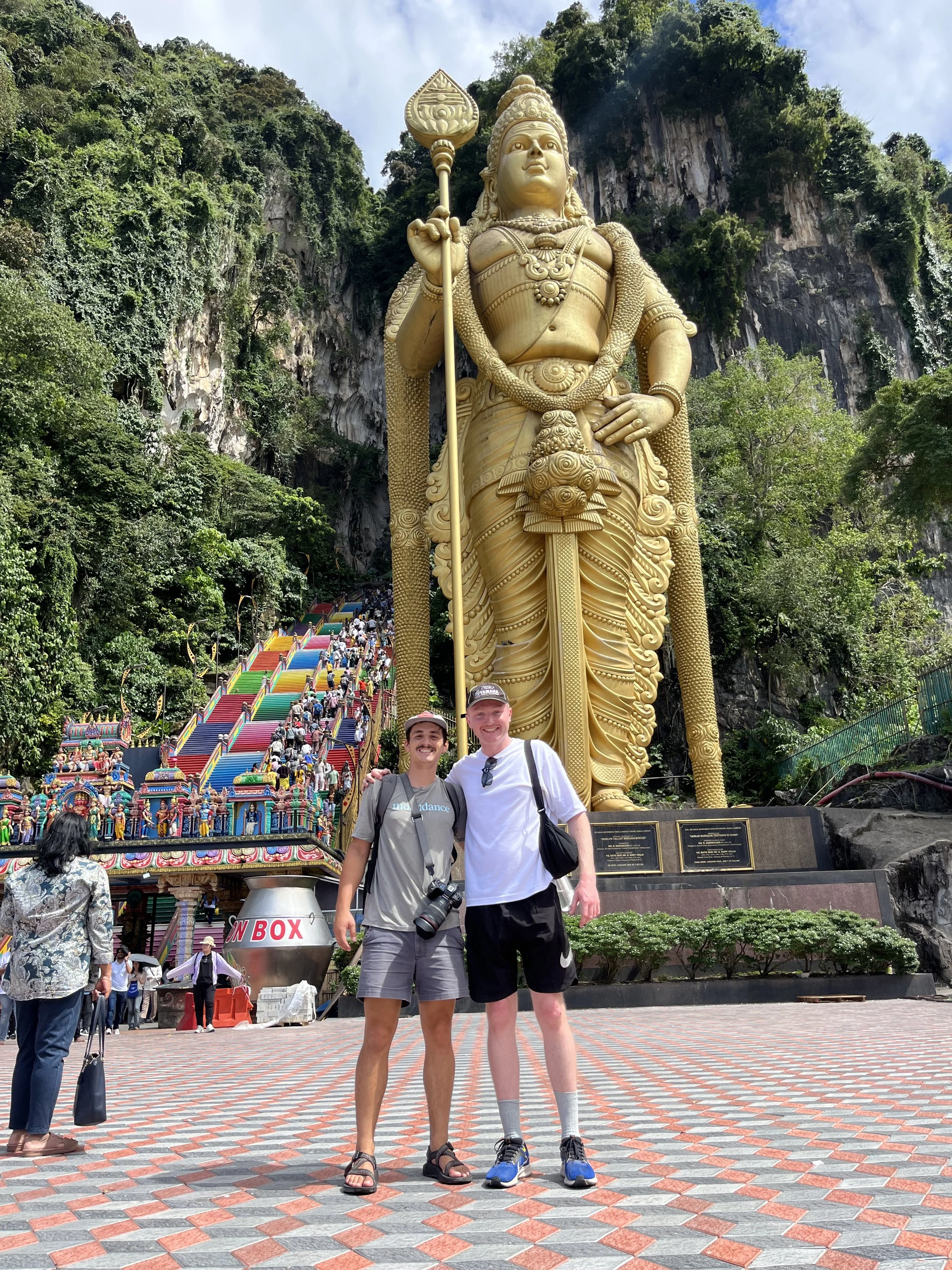 Two travelers standing in front of the golden Murugan statue at Batu Caves in Kuala Lumpur, with the colorful staircase and limestone cliffs rising behind them.