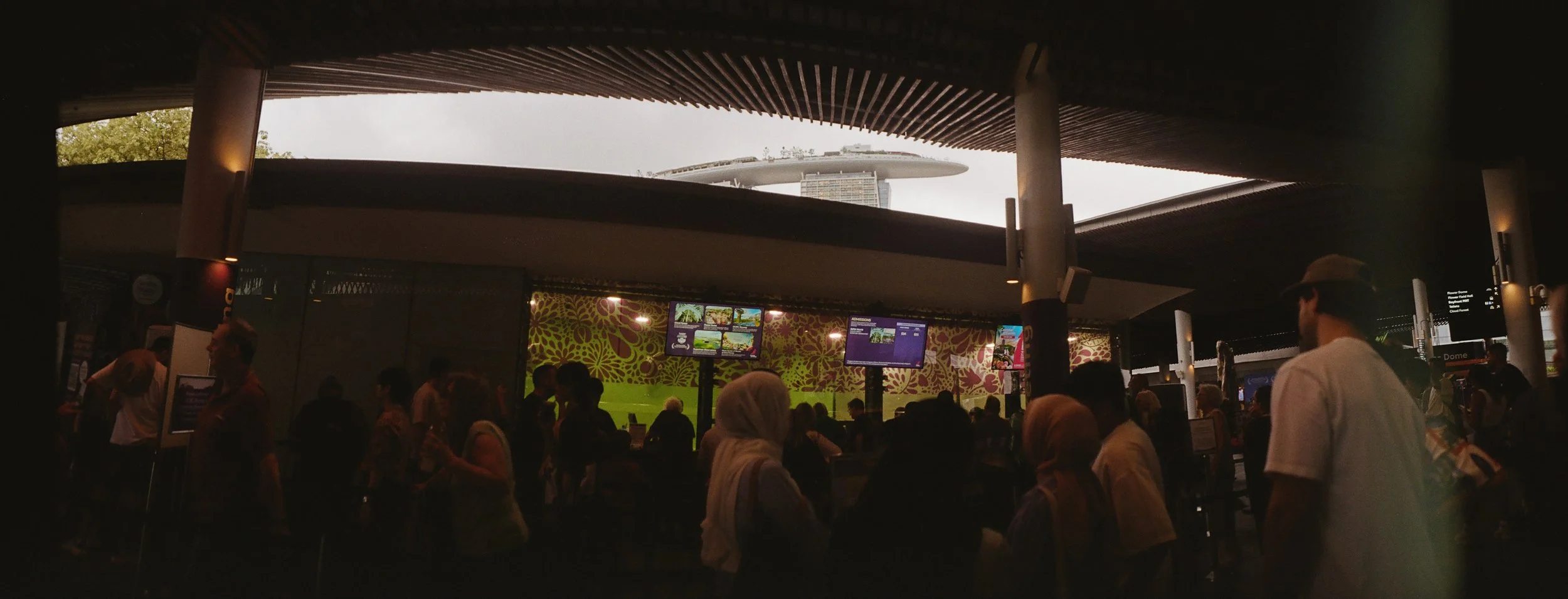 Crowds gathered under the open-air structure at Gardens by the Bay in Singapore, with Marina Bay Sands visible through the curved roof above.