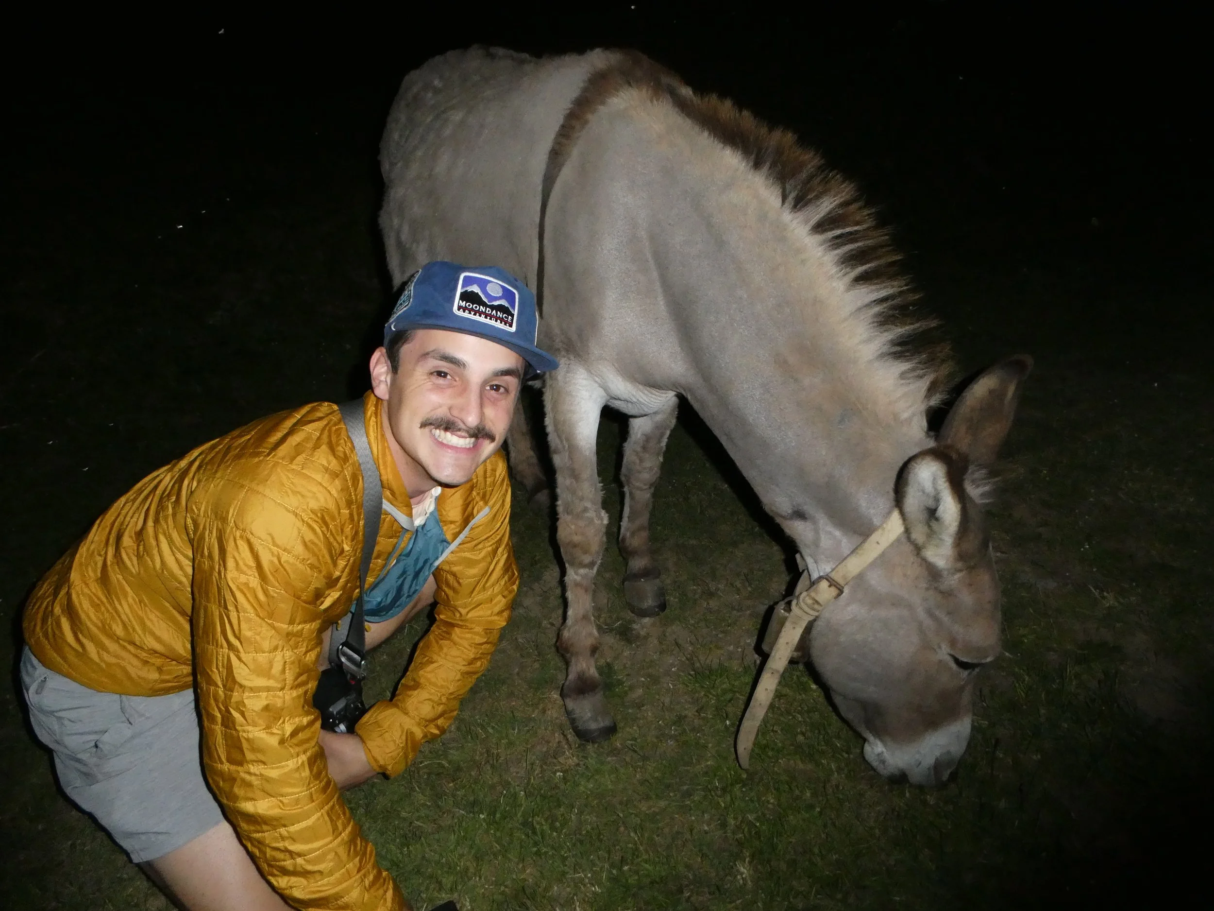 Sebastian crouching next to a light brown donkey grazing on grass at night, smiling at the camera in northern spain, Vegabano Refugio.