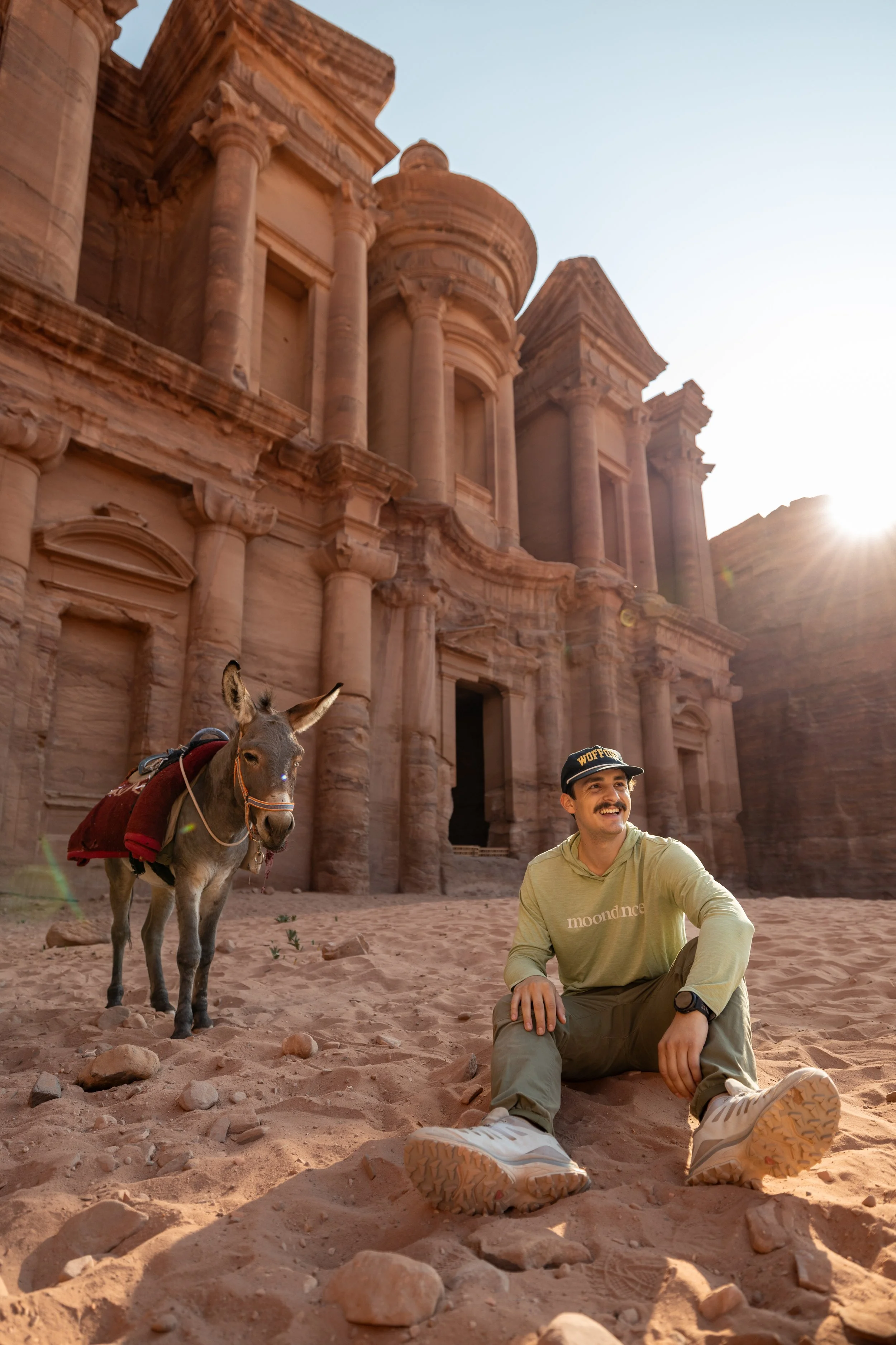 Sebastian sitting with a donkey in front of an ancient sandstone building with tall columns and intricate carvings, during sunset.