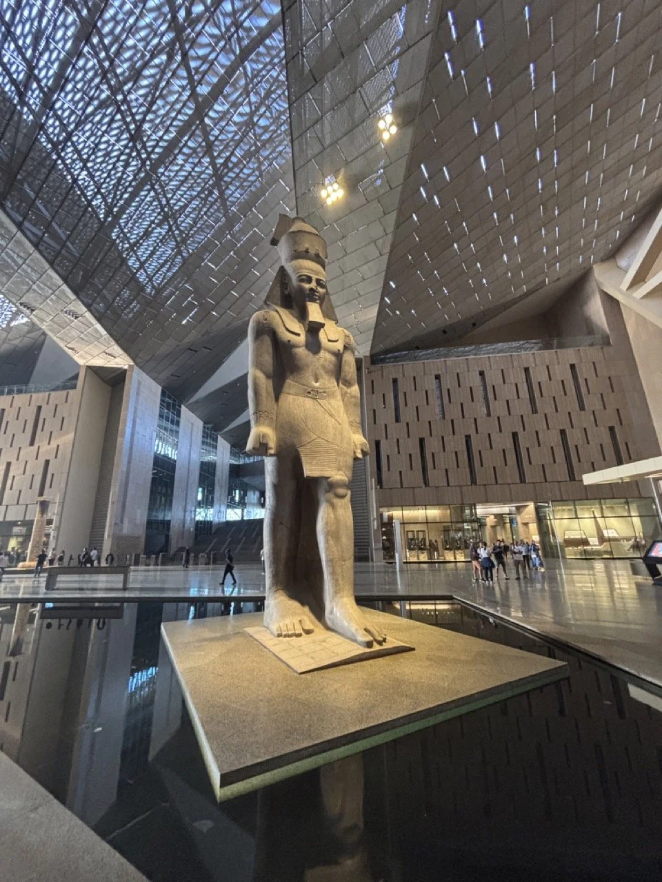 Colossal ancient Egyptian statue displayed inside the Grand Egyptian Museum in Cairo, reflected in polished stone floors beneath the museum’s dramatic modern architecture.