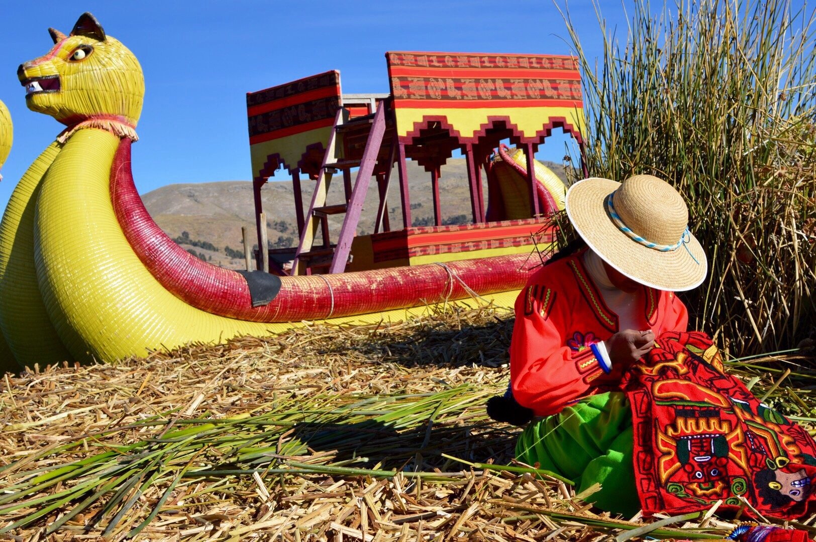 Indigenous woman wearing traditional clothing sewing colorful textiles beside a reed boat on the floating islands of Lake Titicaca in Peru