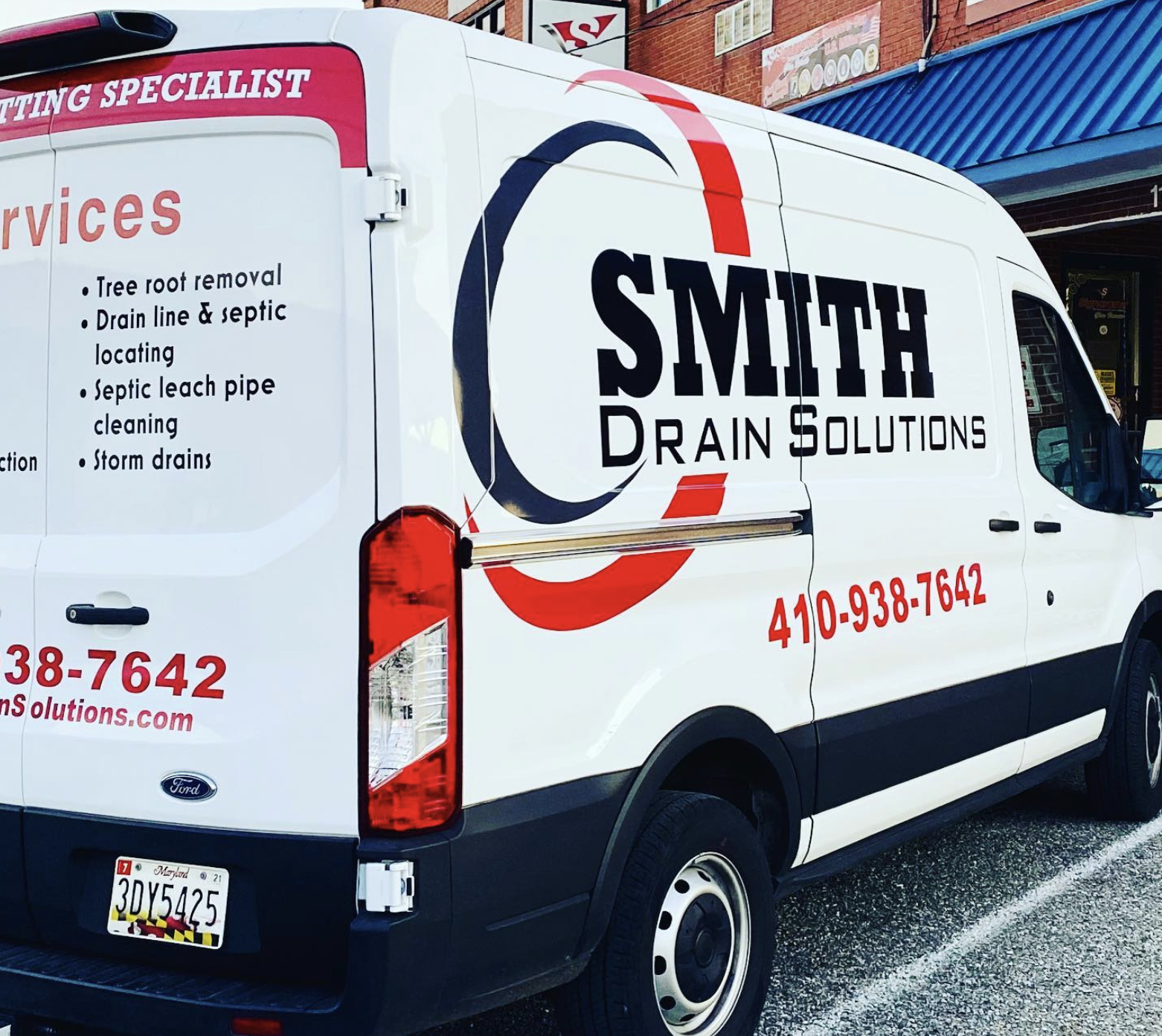 A white Ford service van for Smith Drain Solutions parked outside a building. The van displays services like tree root removal, drain line and septic locating, septic leach pipe cleaning, and storm drains, along with the company's phone number 410-938-7642 and website.