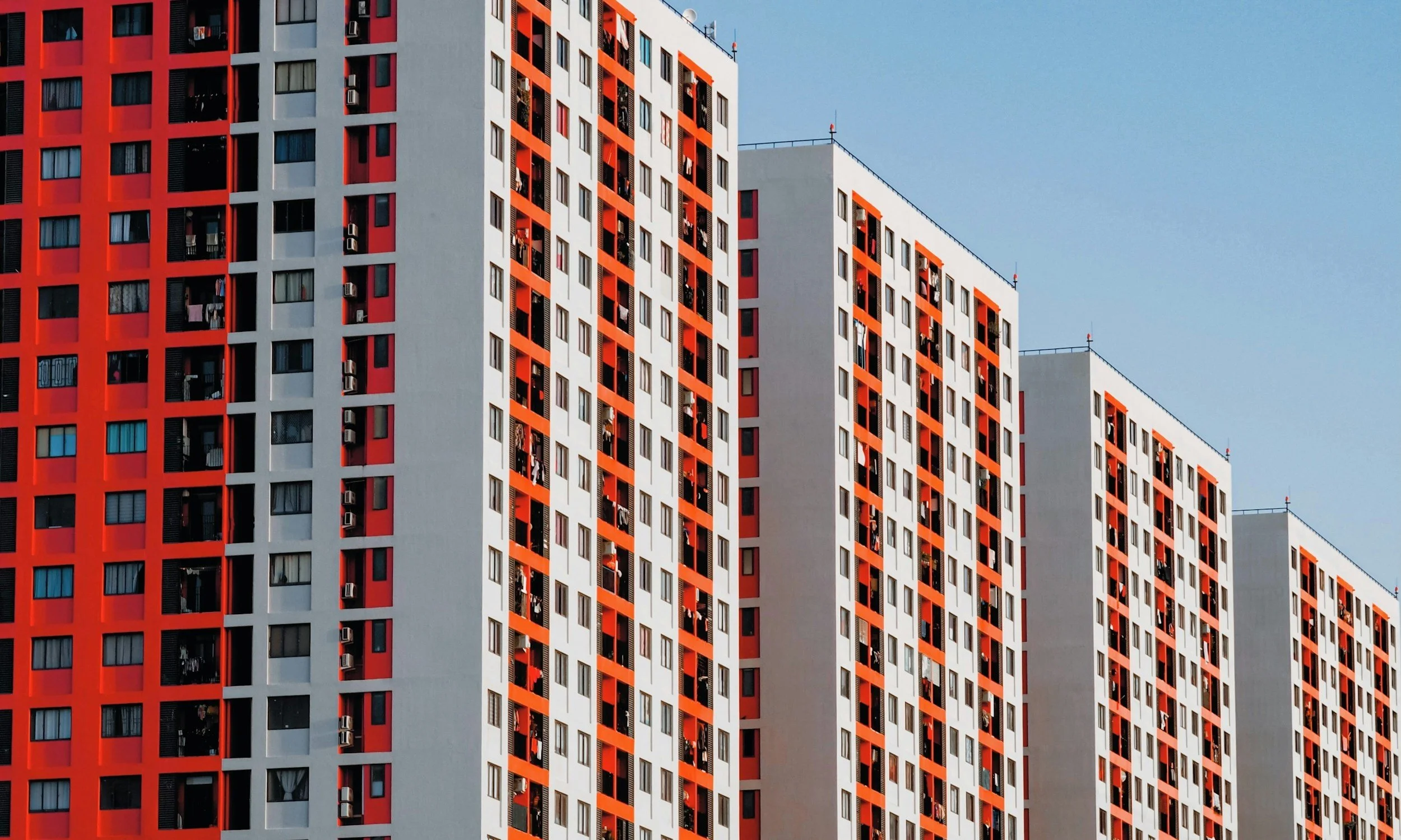 High-rise apartment building with alternating red and white sections, numerous windows and balconies, and a clear blue sky in the background.