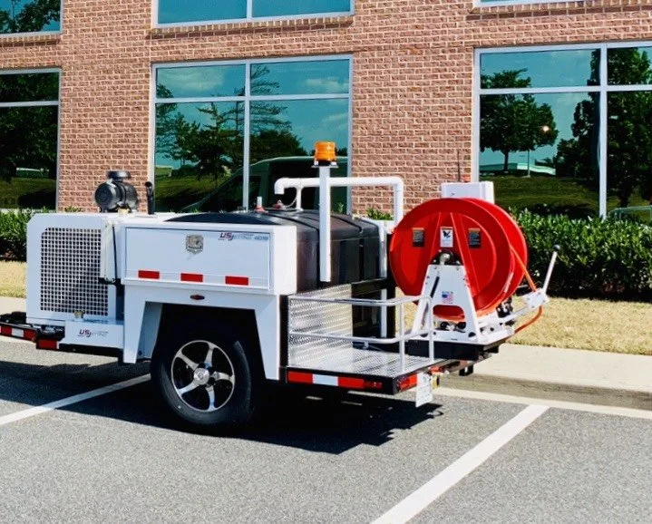 Fire truck service vehicle with a red hose reel, parked in a parking lot outside a brick building with large windows.