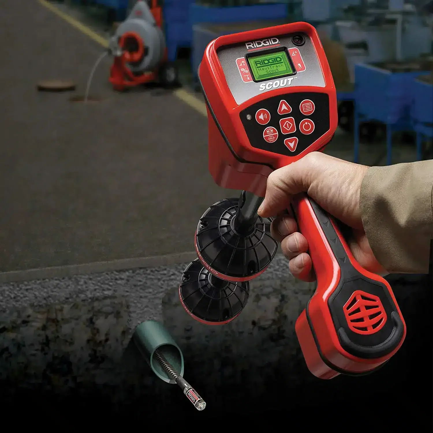A person holding a red RIDGID electronic pipe inspection camera with a monitor and controls. The camera appears to be inserted into a pipe below ground level.