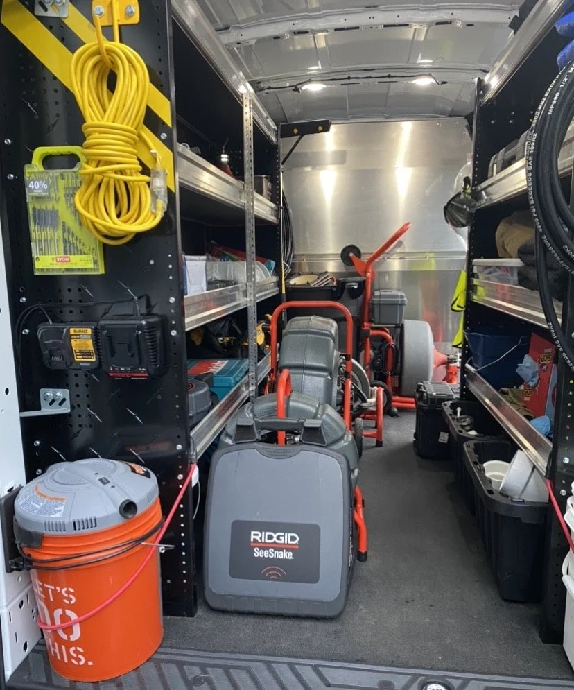 Interior of a utility truck with various tools and equipment. Includes yellow extension cord, black power drill with yellow label, orange vacuum cleaner labeled 'RIDGID SeeSnake', shelves with supplies, and black storage containers.